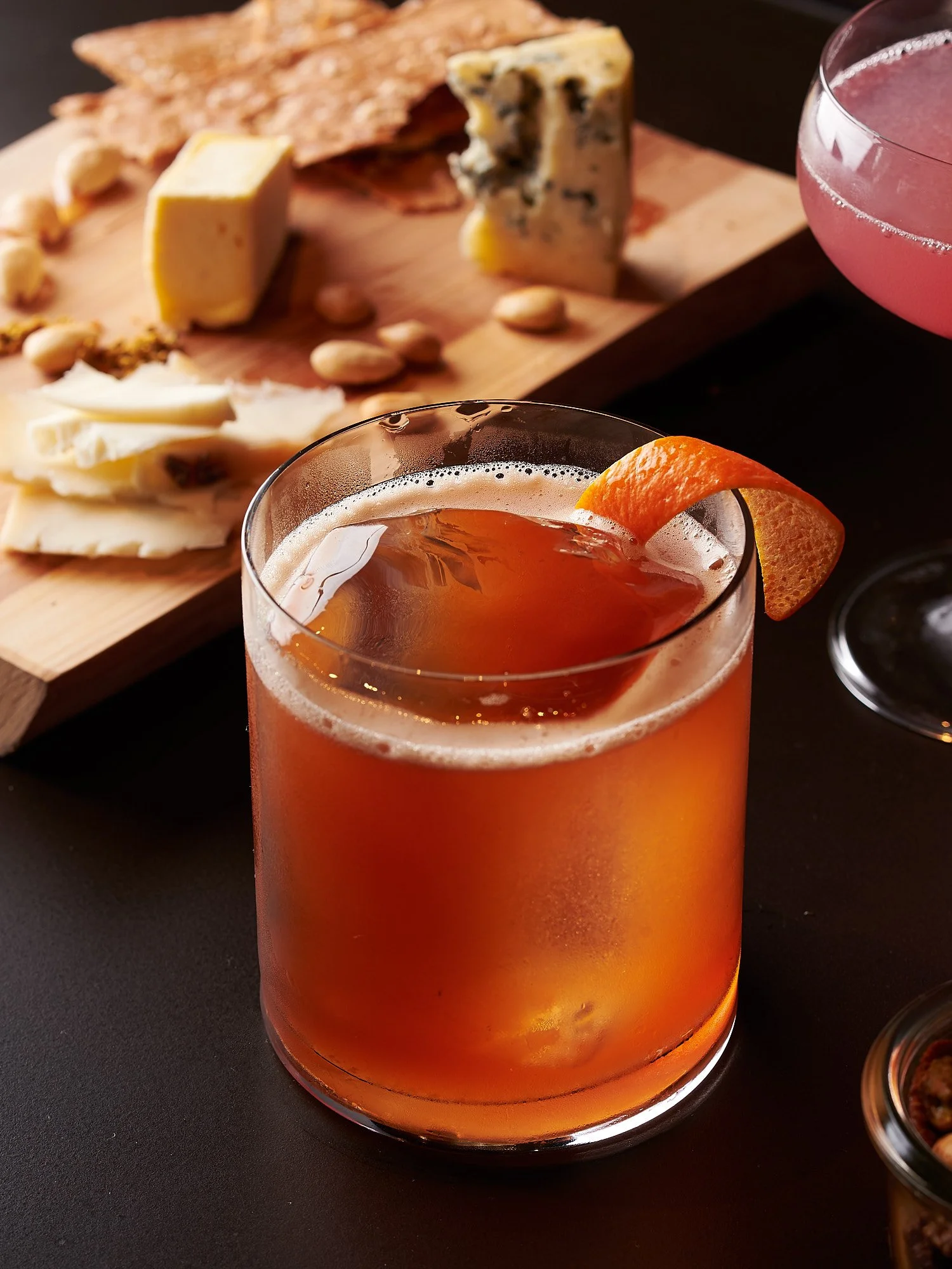 Beverage advertising photography by Dhanraj Emanuel — amber cocktail with orange twist garnish alongside cheese and charcuterie board. Commercial drink photographer for spirits brands and restaurant clients, available nationwide.