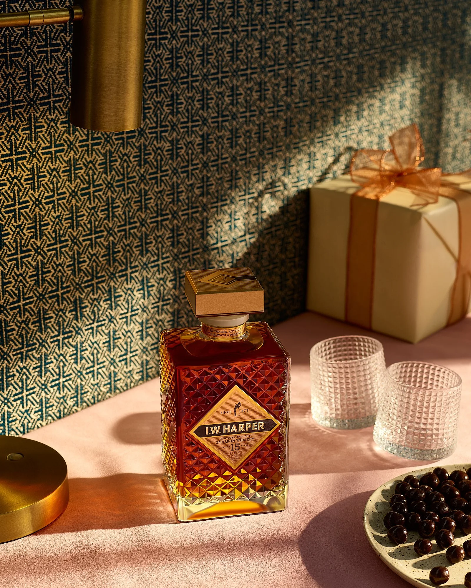 Front view of a bottle of I.W. Harper bourbon whiskey on a pink table, next to two empty textured glass cups, a plate of dark cherries, a wrapped gift, and a gold lamp base. The background features blue and gold patterned wallpaper.