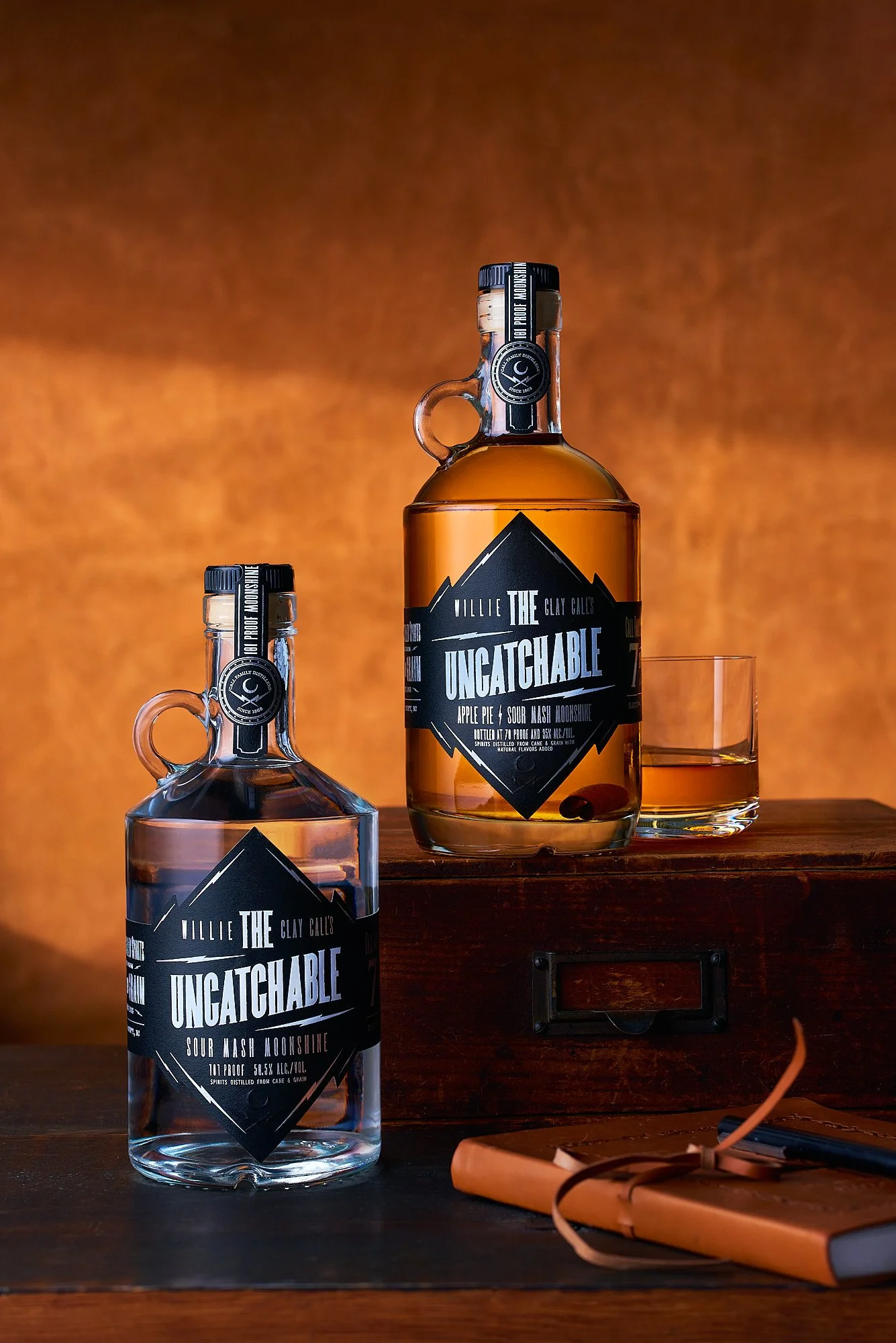Spirits advertising photography by Dhanraj Emanuel — Call Family Distillers whiskey and moonshine bottles styled with leather notebook and wooden box on dark surface. Commercial beverage photographer for craft spirits brands, available nationwide.