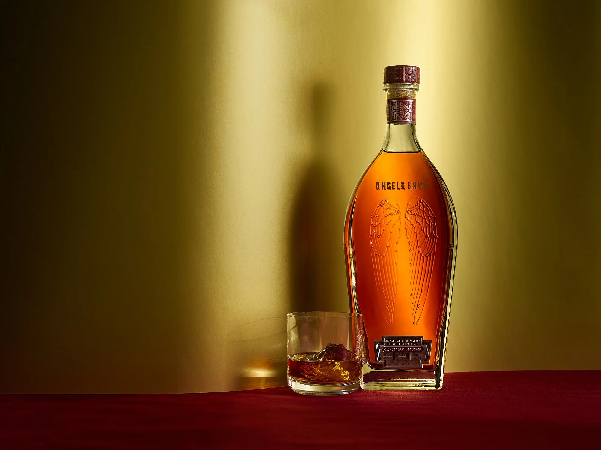 A bottle of Angel's Envy bourbon whiskey with an engraved angel wing design and a glass of bourbon with ice on a red surface against a yellow backdrop.