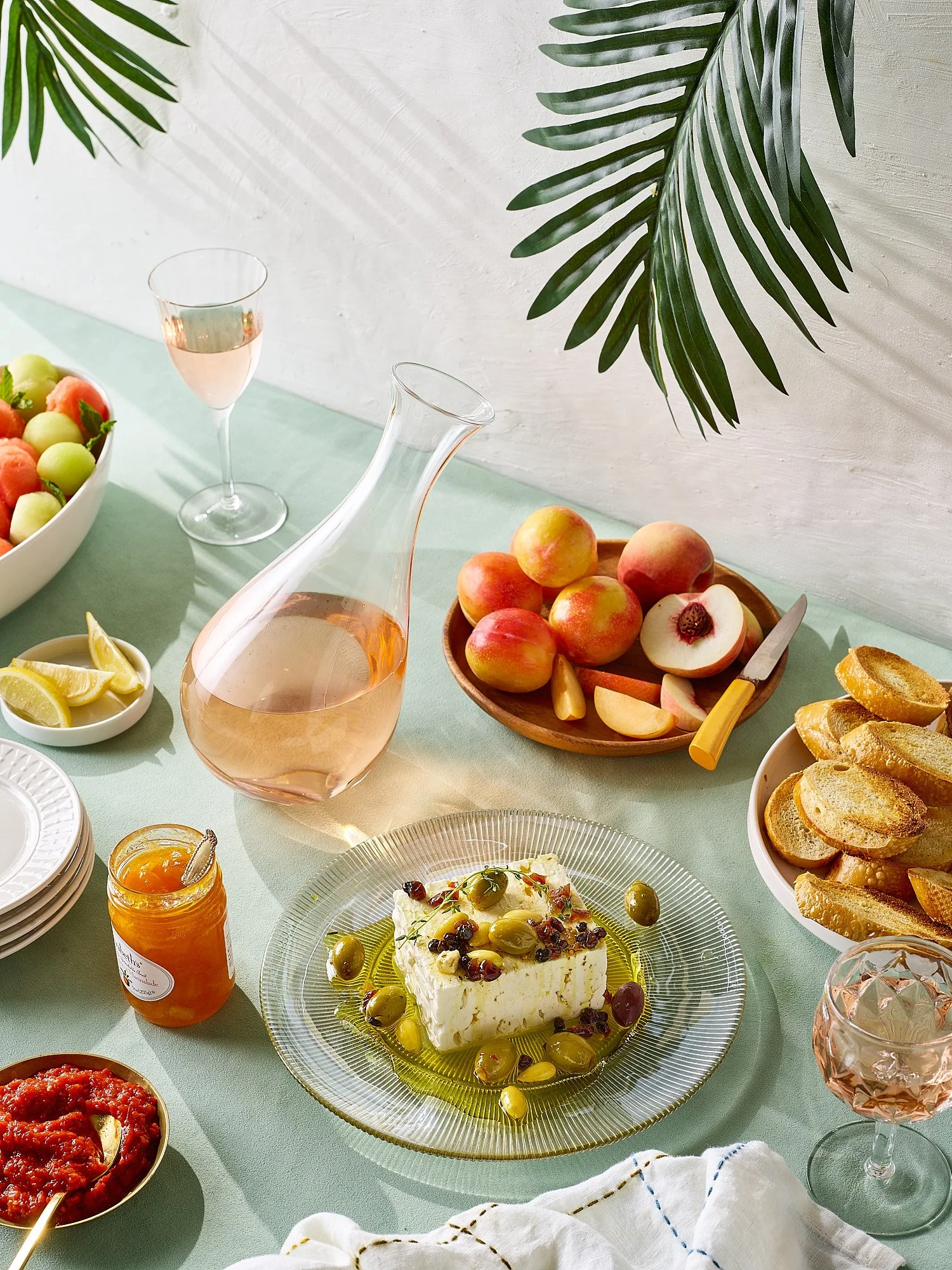 A table set with various foods including a bowl of fruit, a carafe of rosé wine, a glass of rosé, a plate of cheese with olives and herbs, a bowl of toasted slices of bread, a small jar of apricot jam, lemon wedges, and a bowl of tomato-based sauce w