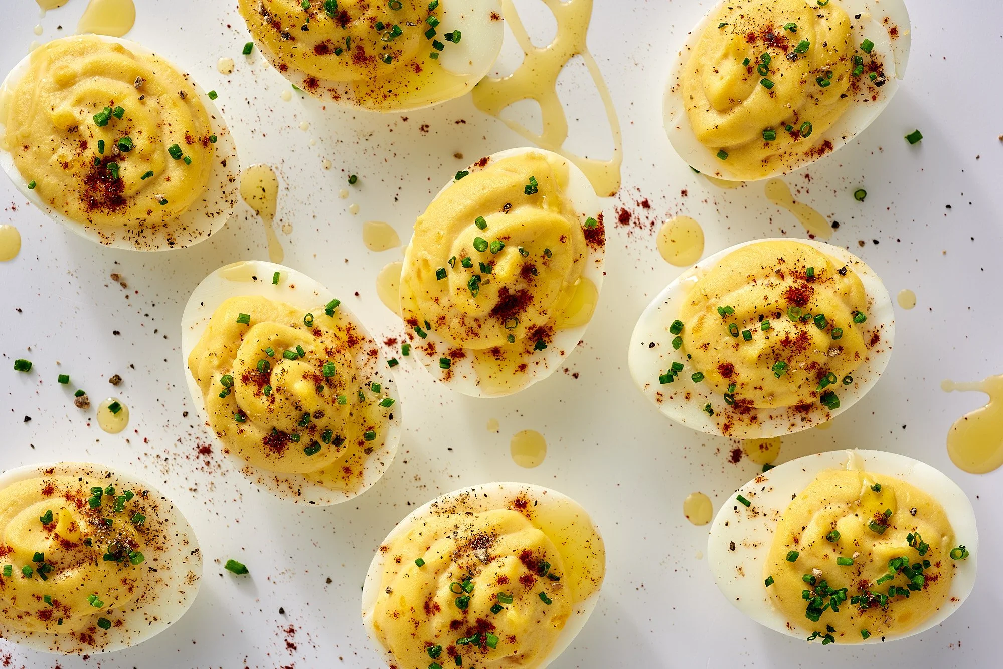Deviled eggs topped with chopped chives, paprika, and drizzled with olive oil on a white surface.
