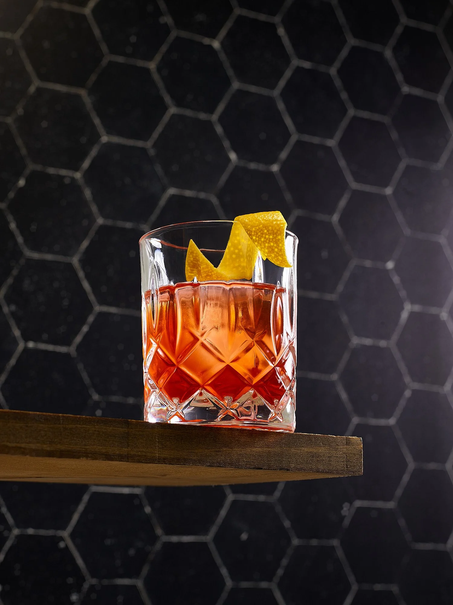 A glass of red cocktail garnished with lemon peel, on a wooden surface with a black honeycomb-patterned background.