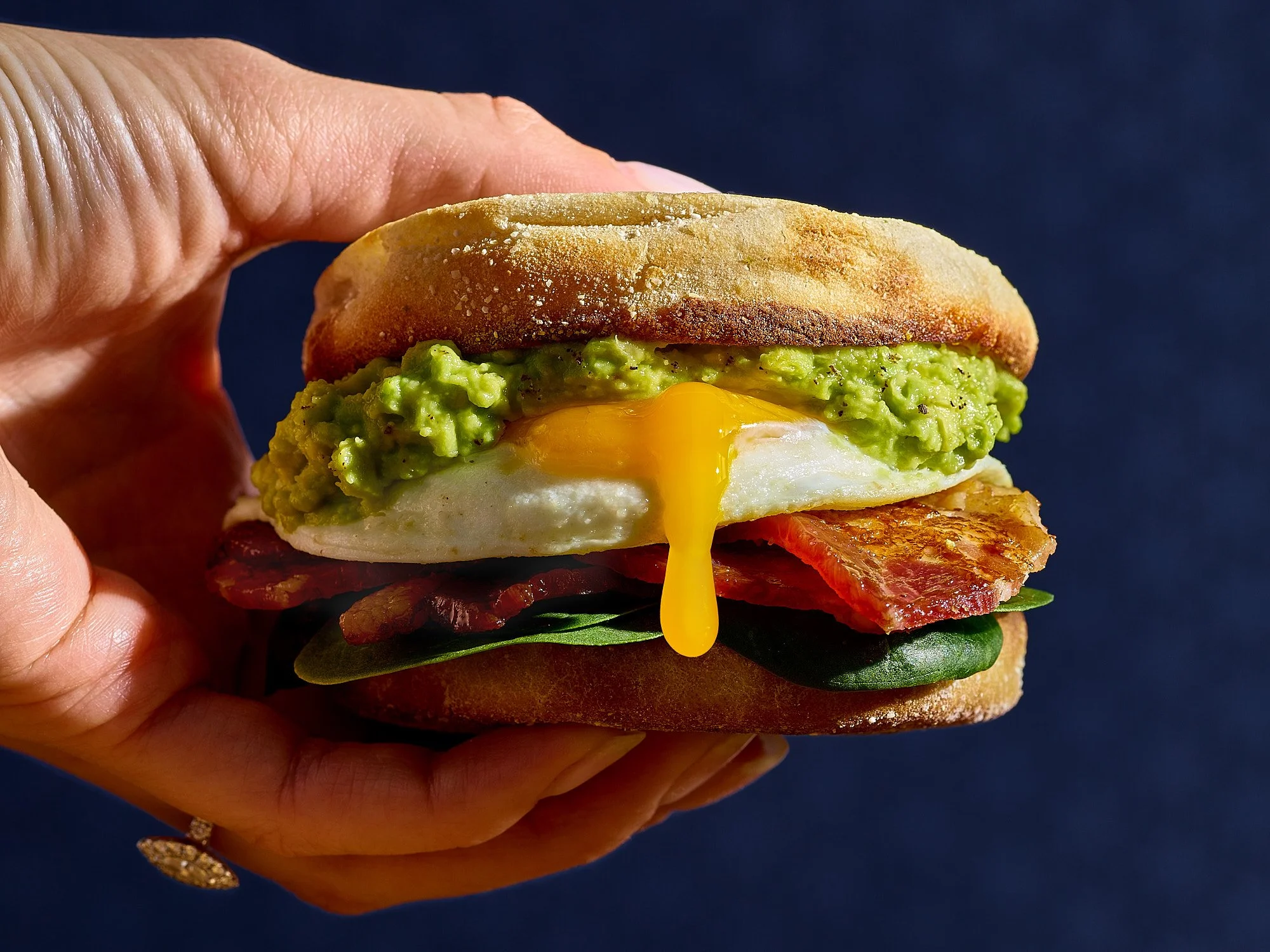 Food advertising photography by Dhanraj Emanuel — hand-held breakfast sandwich with fried egg, avocado, and crispy bacon on toasted English muffin. CPG and restaurant food photographer based in Greensboro NC.