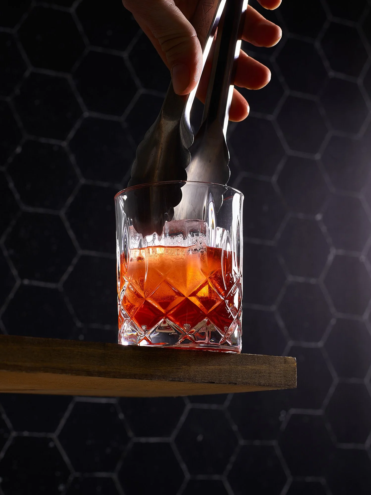 Spirits advertising photography by Dhanraj Emanuel — hand placing large ice cube into amber whiskey with metal tongs, dark dramatic background. Commercial beverage photographer for spirits brands and advertising agencies, available nationwide
