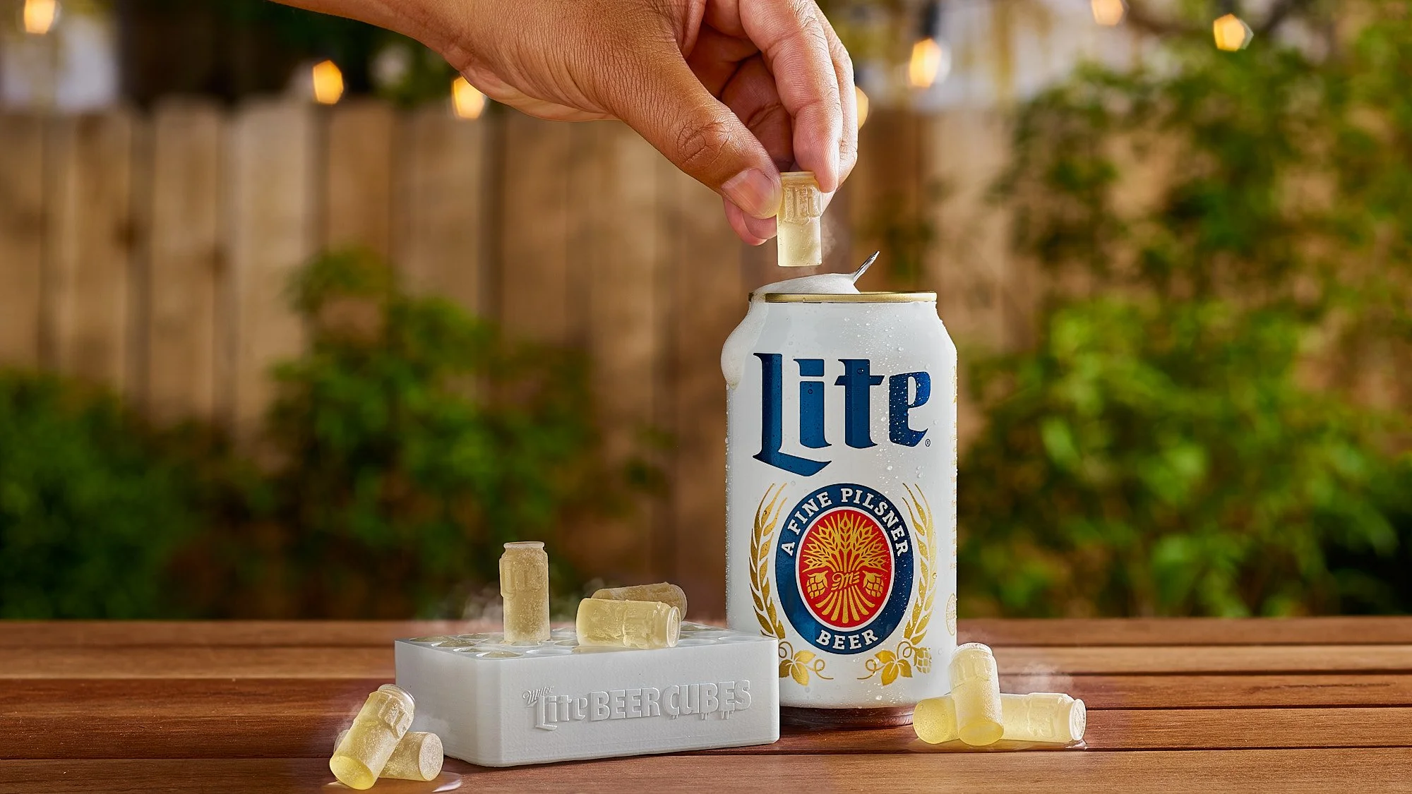 A hand dropping a beer cube into a can of Miller Lite beer outdoors. Several beer cubes are scattered on a wooden surface, with a white Miller Lite beer cube holder nearby.
