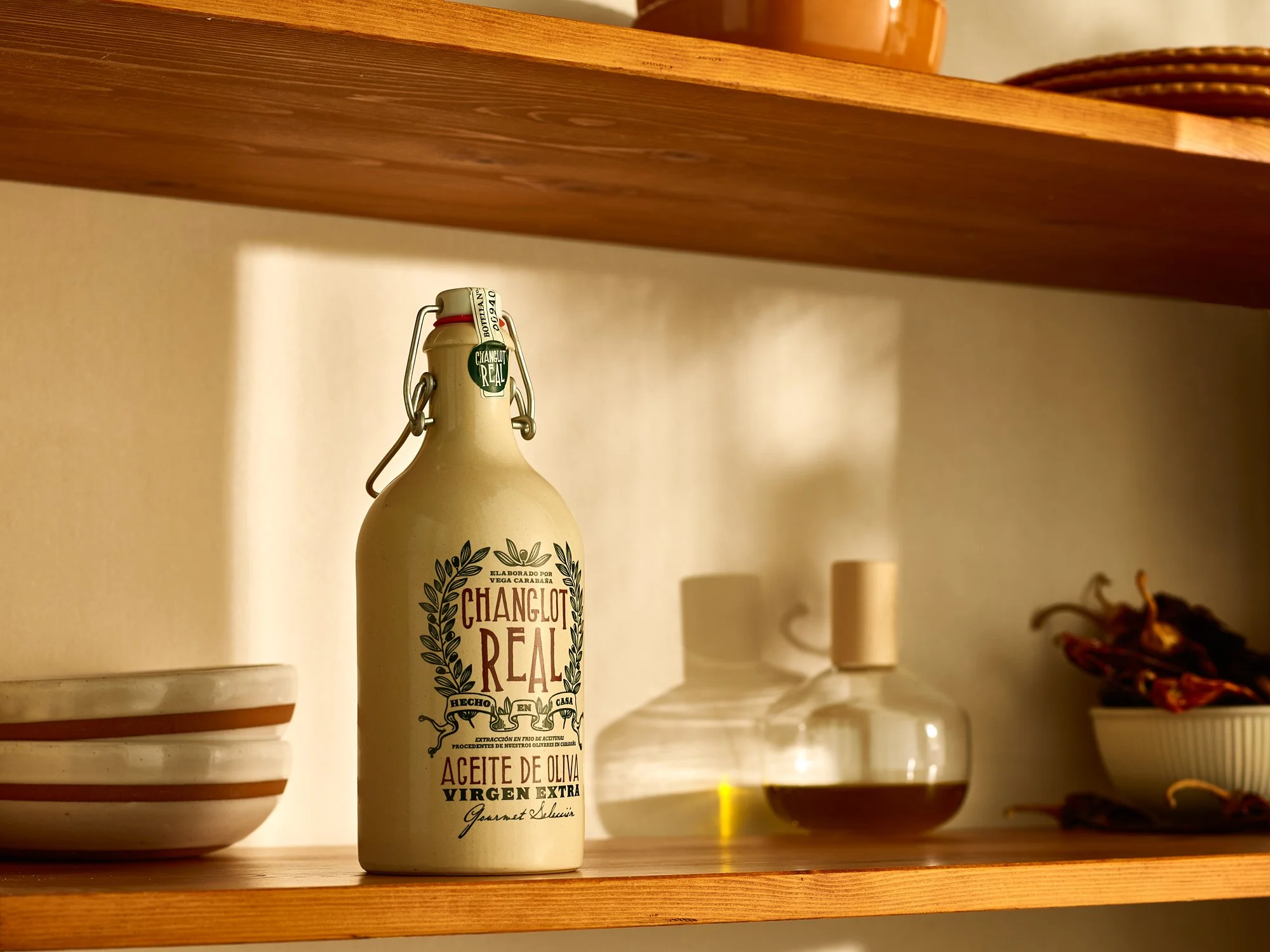Changlot Real extra virgin olive oil in a stoneware swing-top bottle, styled on a warm wooden kitchen shelf beside ceramic bowls, a glass cruet of oil, and dried peppers. Food and beverage advertising photography by Dhanraj Emanuel.