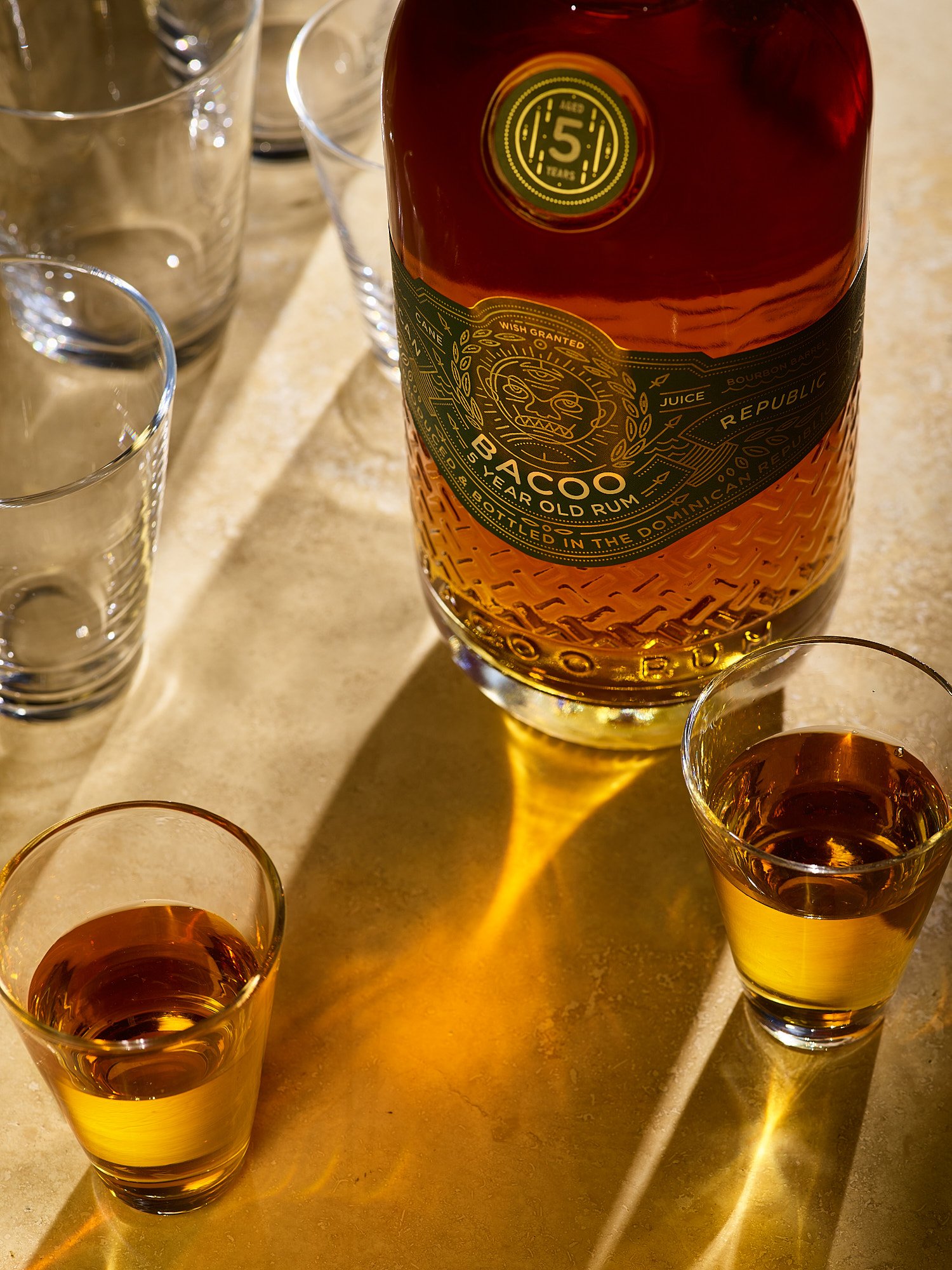 Beverage advertising photography by Dhanraj Emanuel — Bacoo 5-Year-Old Rum bottle with styled glasses on light surface. Commercial spirits photographer for rum and beverage brands, working nationally for agencies and brands.