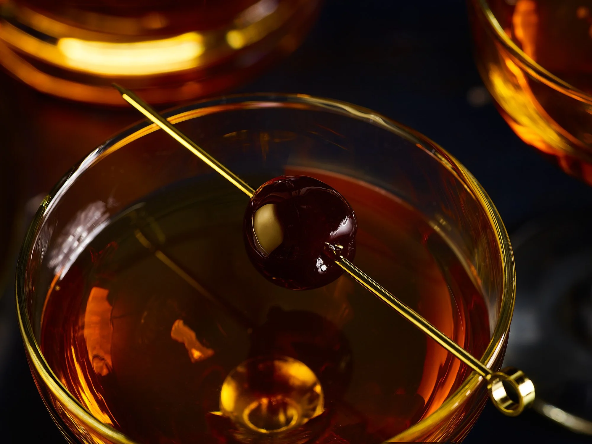 Close-up of a cocktail glass with dark liquid, garnished with cherries on a cocktail pick.