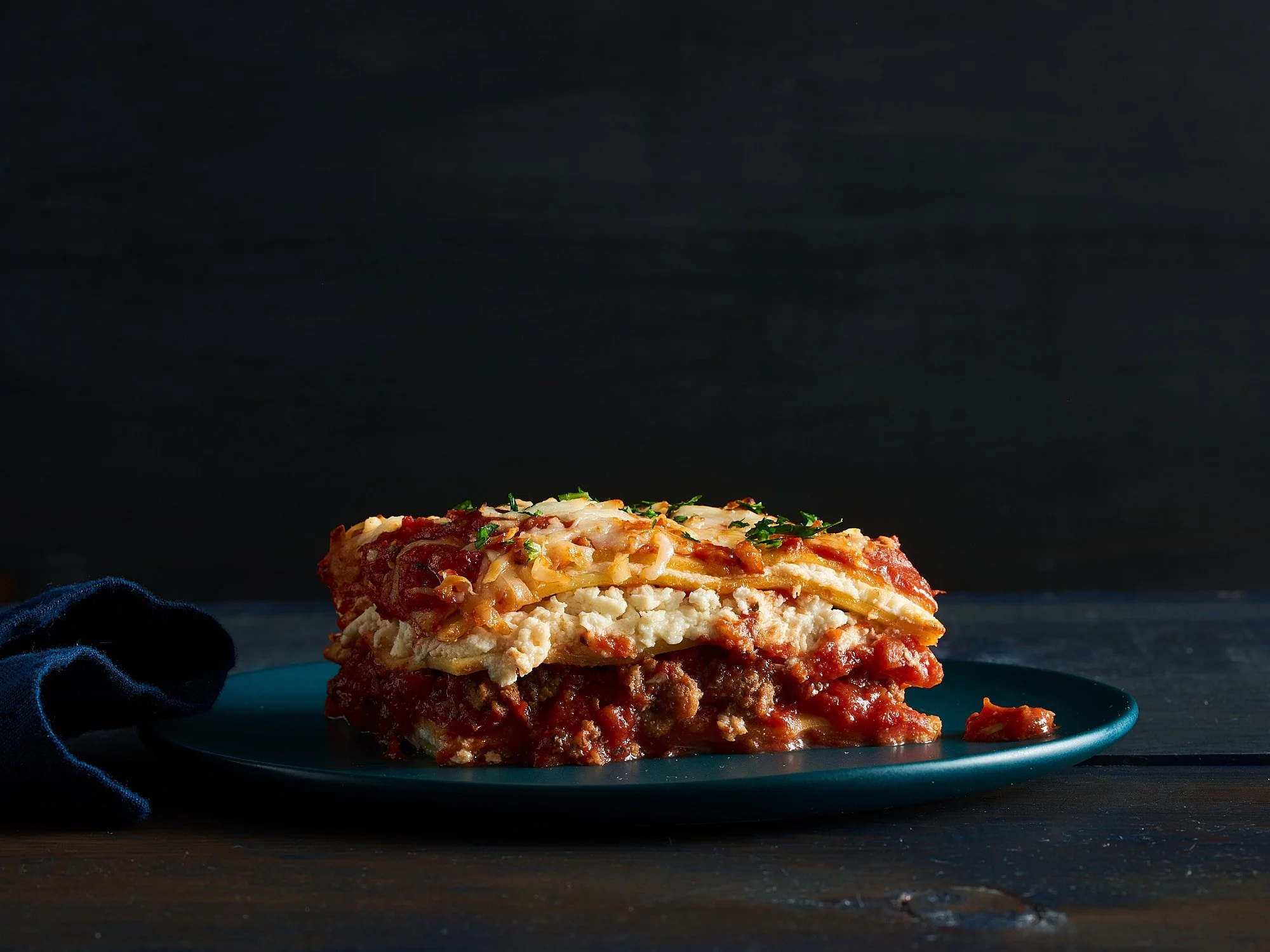 Food advertising photography by Dhanraj Emanuel — dramatic dark-background lasagna on blue ceramic, layered pasta with meat sauce and herbs. Commercial food photographer for CPG and advertising agencies, available nationwide.