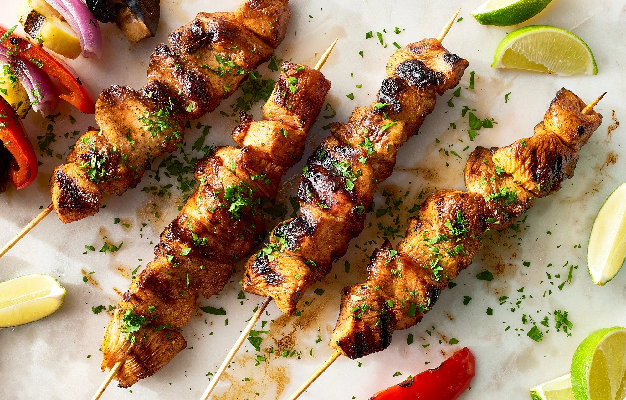 Grilled chicken and beef skewers garnished with chopped herbs, served with lime and lemon wedges and roasted vegetables on a white surface.