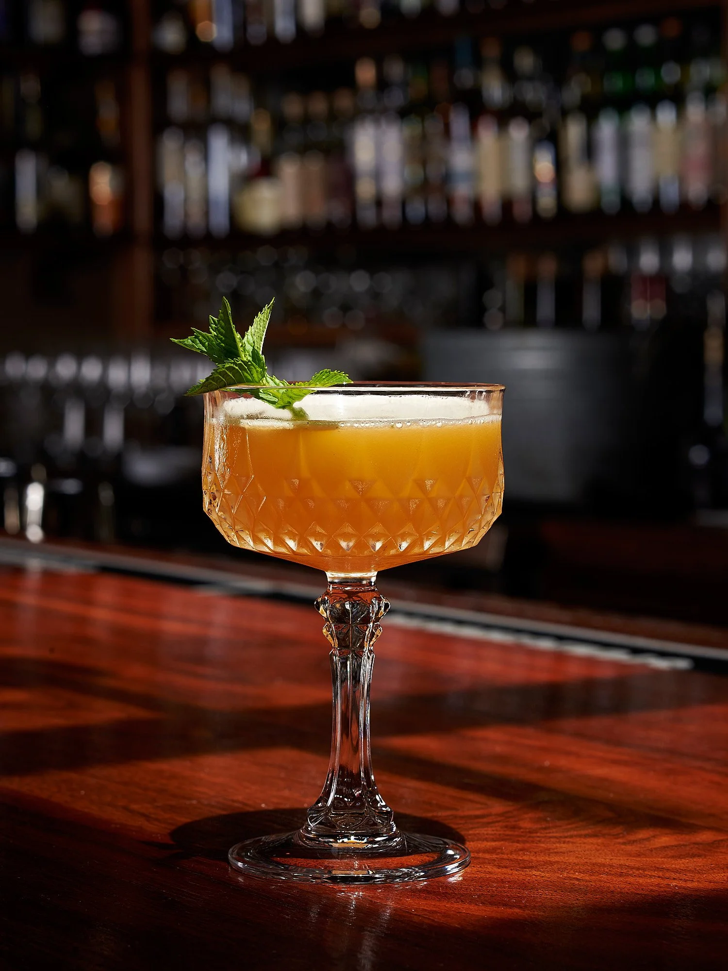 Beverage advertising photography by Dhanraj Emanuel — amber cocktail with mint garnish on vintage glassware at wooden bar counter. Commercial drink photographer for spirits brands, restaurants, and advertising agencies, available nationwide.