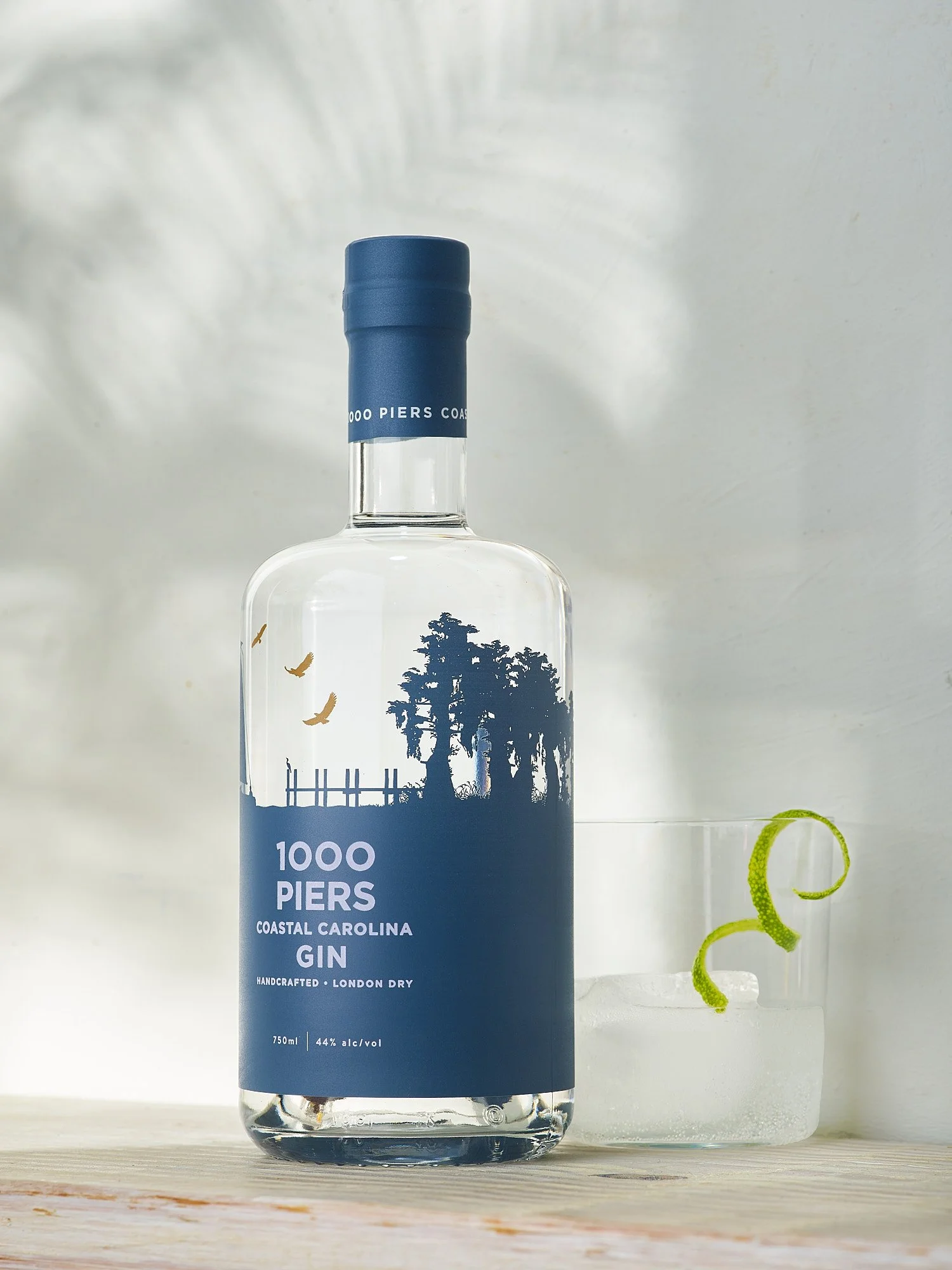 Spirits advertising photography by Dhanraj Emanuel — 1000 Piers Coastal Carolina Gin bottle with lime twist cocktail on wooden surface. Commercial beverage photographer for craft gin and spirits brands, working nationally for agencies and brands.