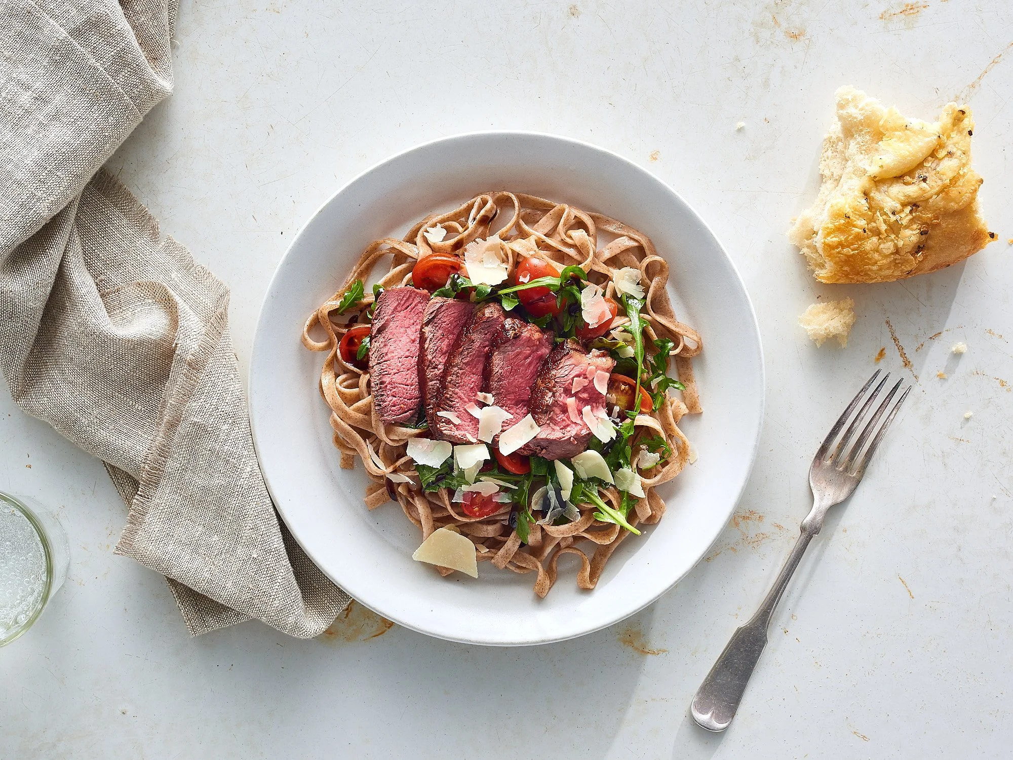 A plate of whole wheat pasta topped with sliced steak, cherry tomatoes, greens, and cheese shavings, with a piece of bread and a fork on a white surface.