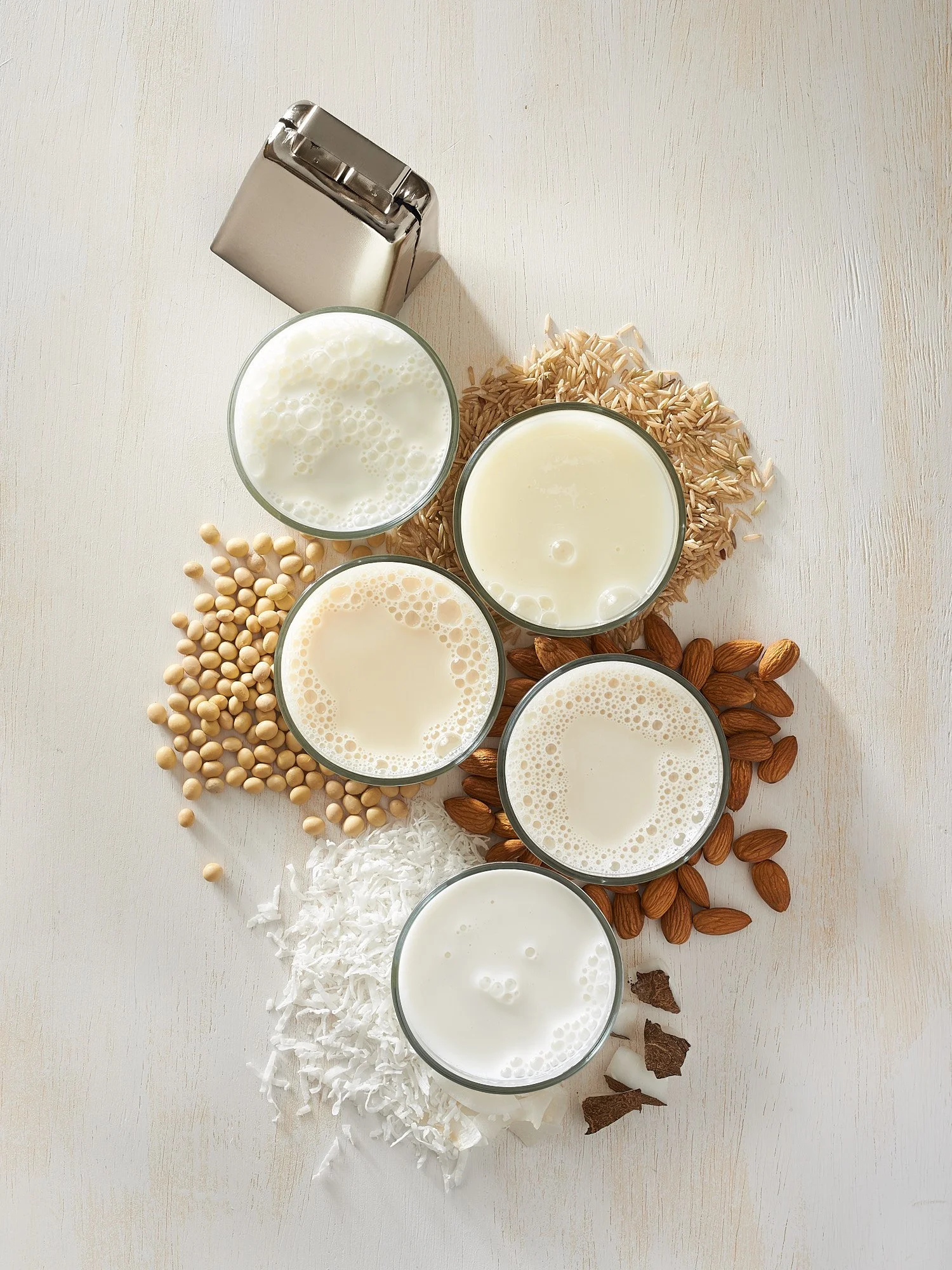 Beverage advertising photography by Dhanraj Emanuel — plant-based nut milks with coconut, grains, and assorted nuts on white surface. Commercial drink photographer for CPG and beverage brands, working nationally for agencies and editorial clients.