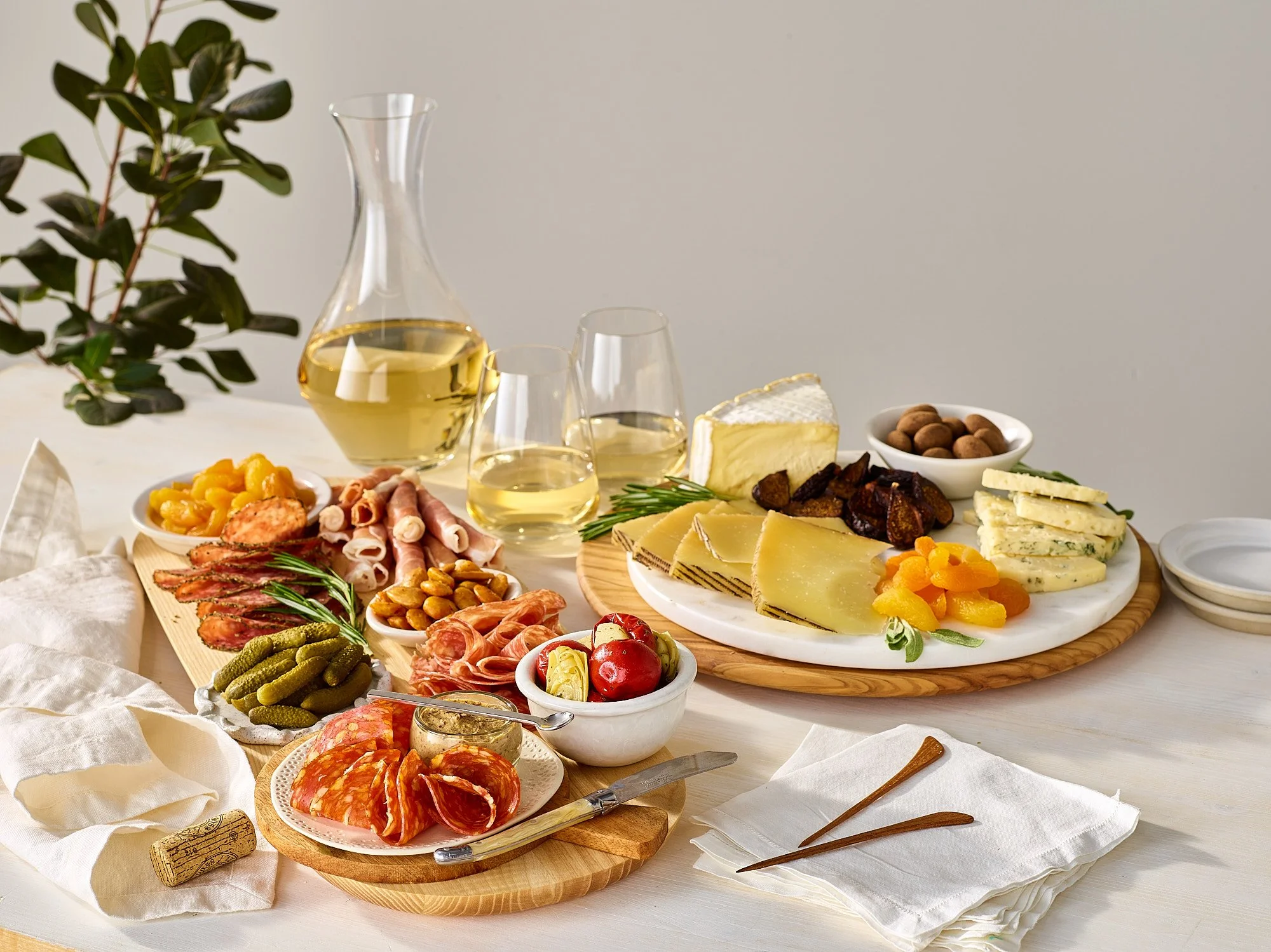 A charcuterie board with cheeses, meats, nuts, and dried fruits, along with a glass of white wine and a decanter, set on a white table with a white backdrop for the fresh market advertising photography by Dhanraj Emanuel, CPG 