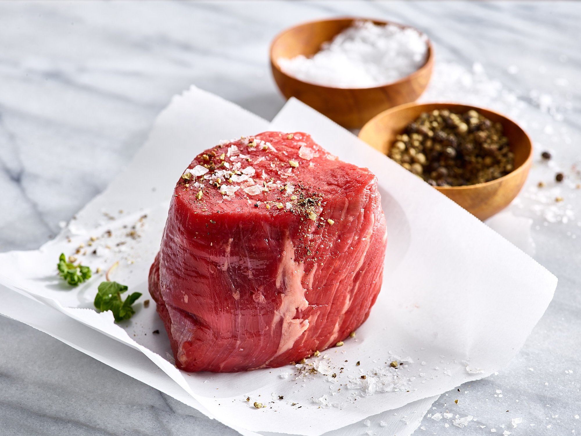 Raw beef steak seasoned with salt and pepper, placed on white parchment paper with a sprig of green herbs, with bowls of salt and peppercorns in the background.