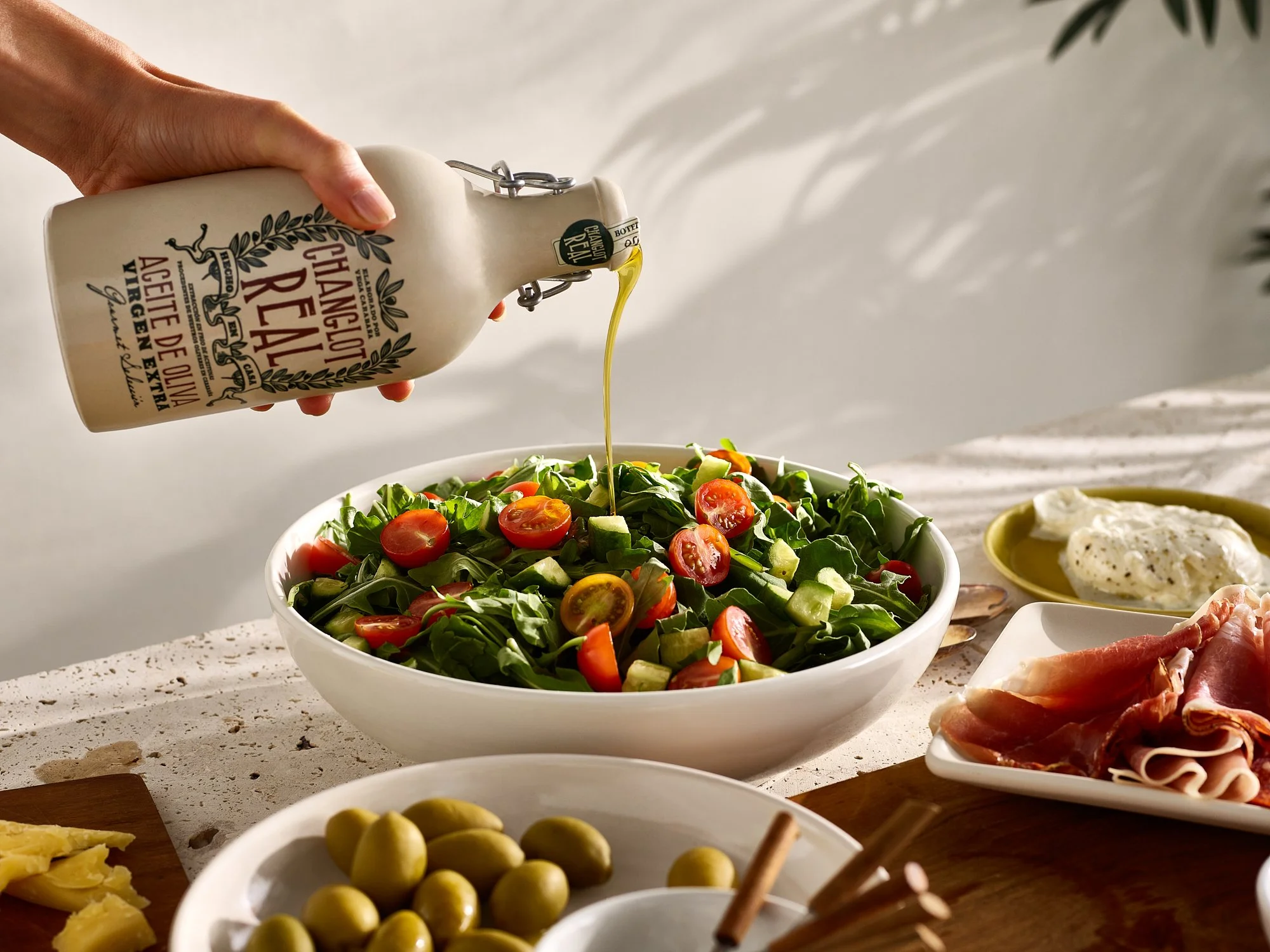 Changlot Real extra virgin olive oil being poured from the stoneware bottle over an arugula and cherry tomato salad, with olives, burrata, and cured meats on the table. Food and beverage advertising photography by Dhanraj Emanuel.
