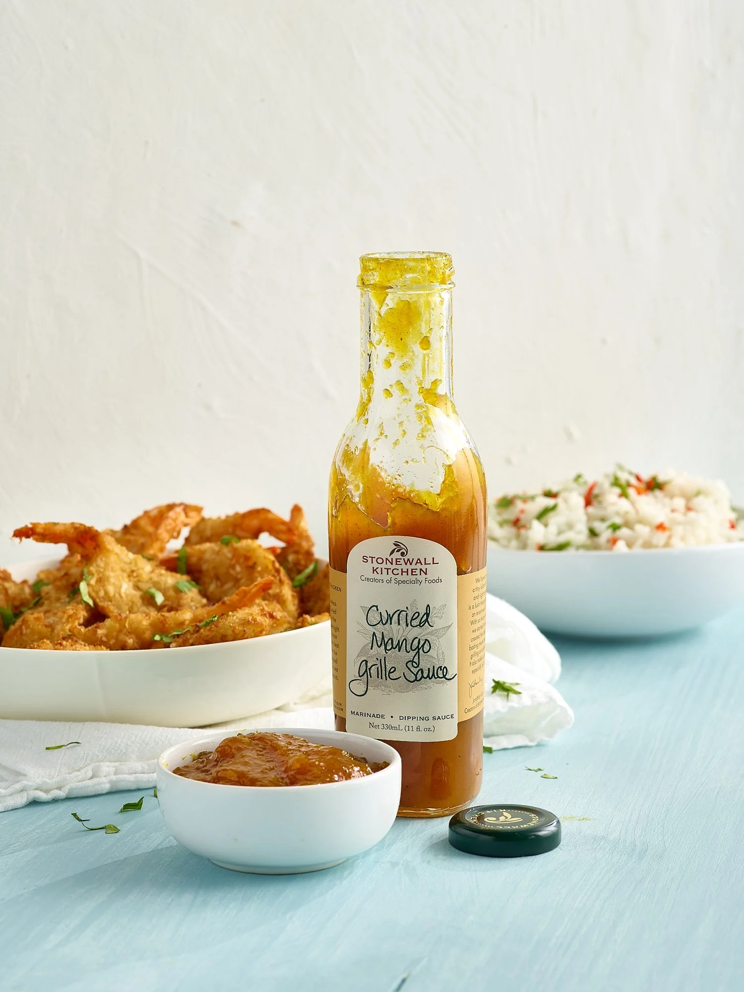 A bottle of Stonewall Kitchen curried mango grill sauce on a light blue surface, with a small bowl of sauce, a bowl of fried food, and a bowl of rice with chopped herbs in the background.