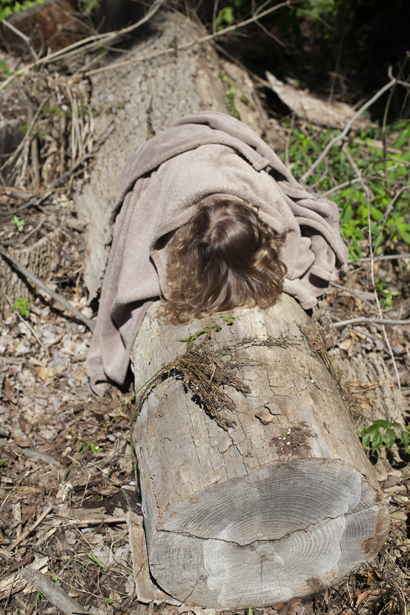 Isa on an Ash Trunk, 2018 In this image, my daughter Isa lies beneath a tan blanket on an Ash log that was felled after being infected by the Emerald Ash Borer. The Ash Borer has dramatically transformed the forest canopy around my childhood home, where Ashes once made up 20% of the tree population. The vulnerability of all living things to environmental changes seems particularly pronounced when looking at these once majestic trunks and their many age rings.
