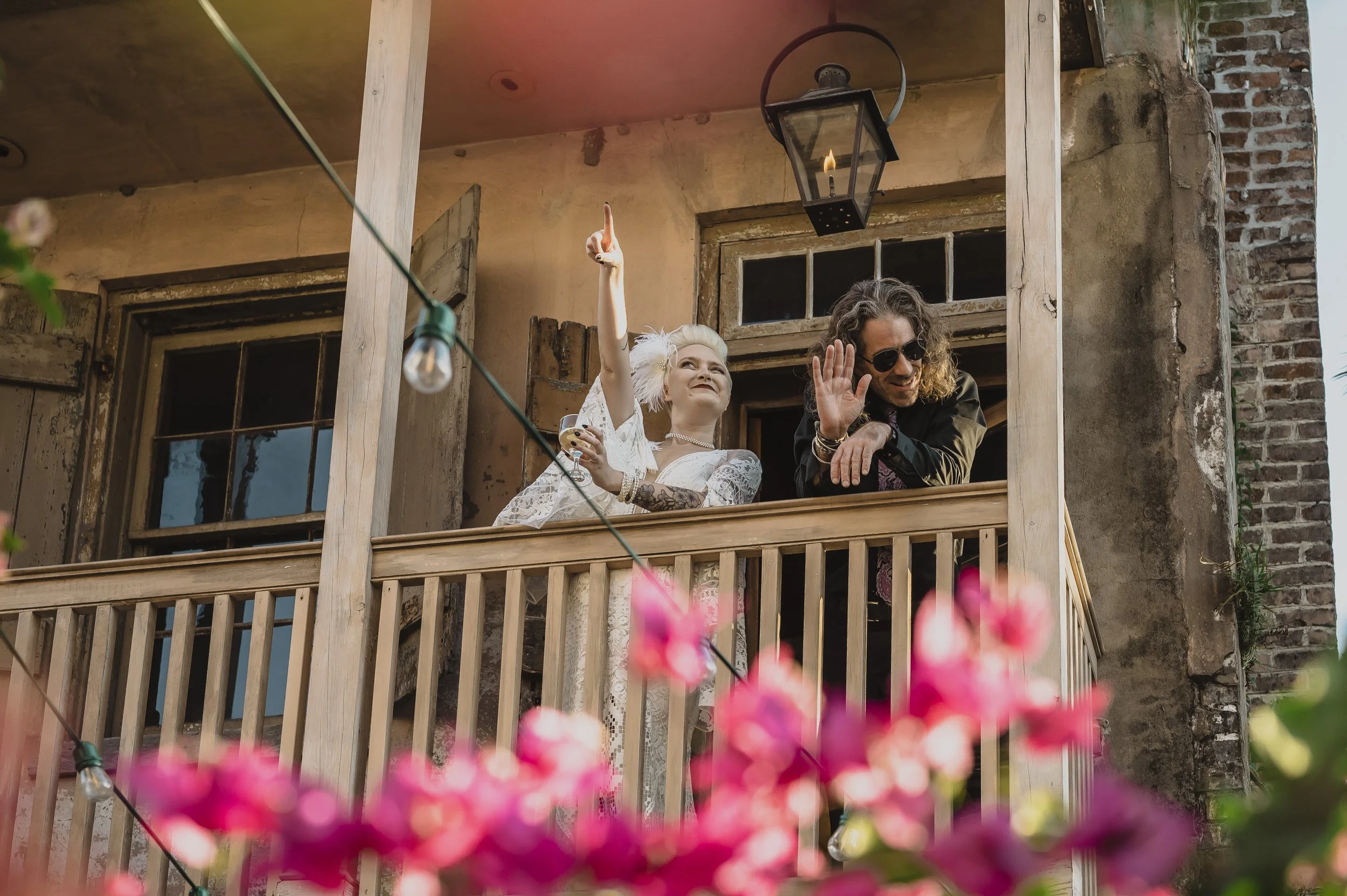 Newlyweds celebrate from a historic New Orleans balcony above bright pink spring flowers.