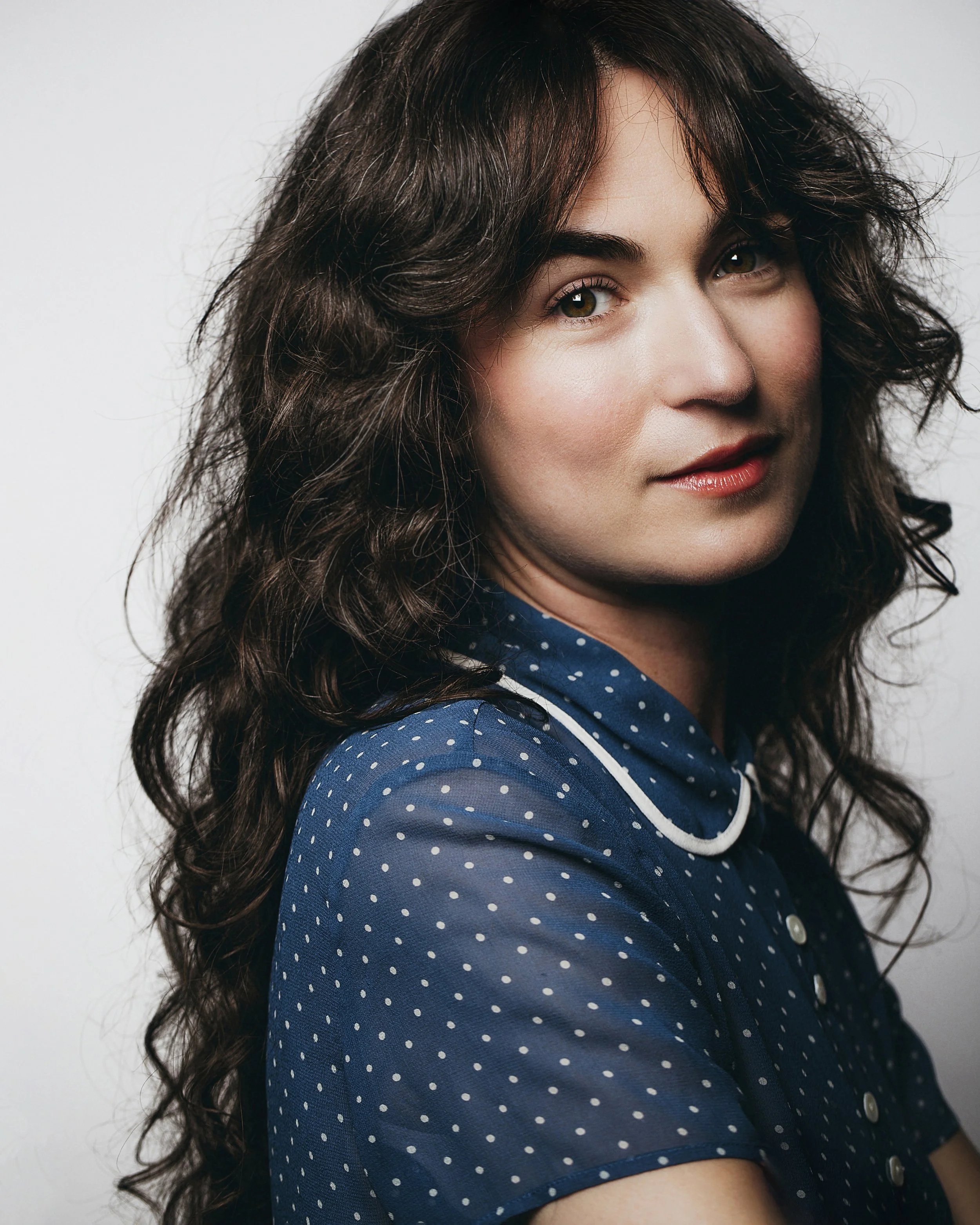 A woman with wavy dark hair and light skin wearing a blue polka dot shirt, looking at the camera with a slight smile.