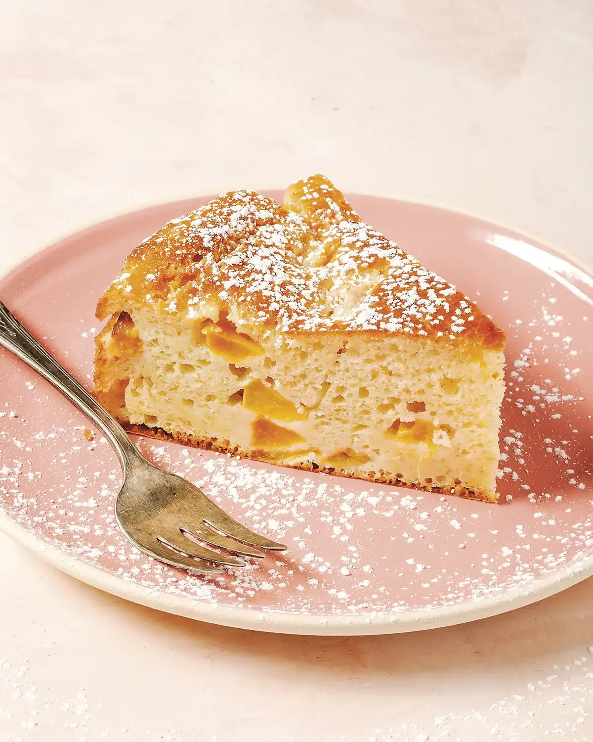 A slice of peach cobbler on a pink plate with powdered sugar, with a fork placed beside it.