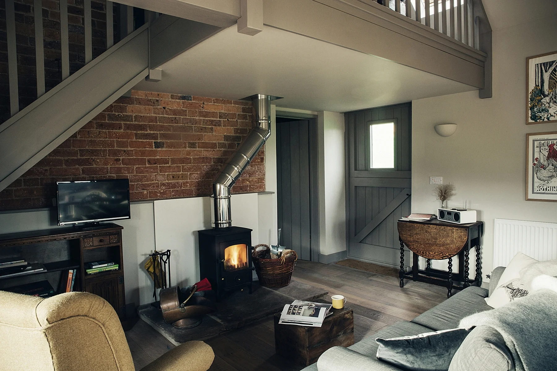 Living room with a wood stove, a yellow armchair, a gray sofa, a small TV on a wooden stand, a coffee table with a magazine and mug, and a brick wall with artwork.