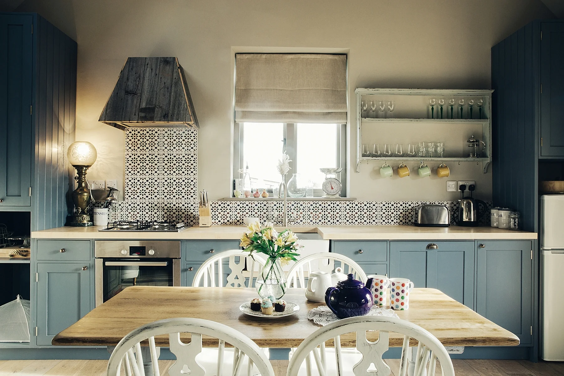 Kitchen with blue cabinets, a wooden table with a vase of flowers, and various kitchen appliances and dishes, illuminated by natural light from a window.