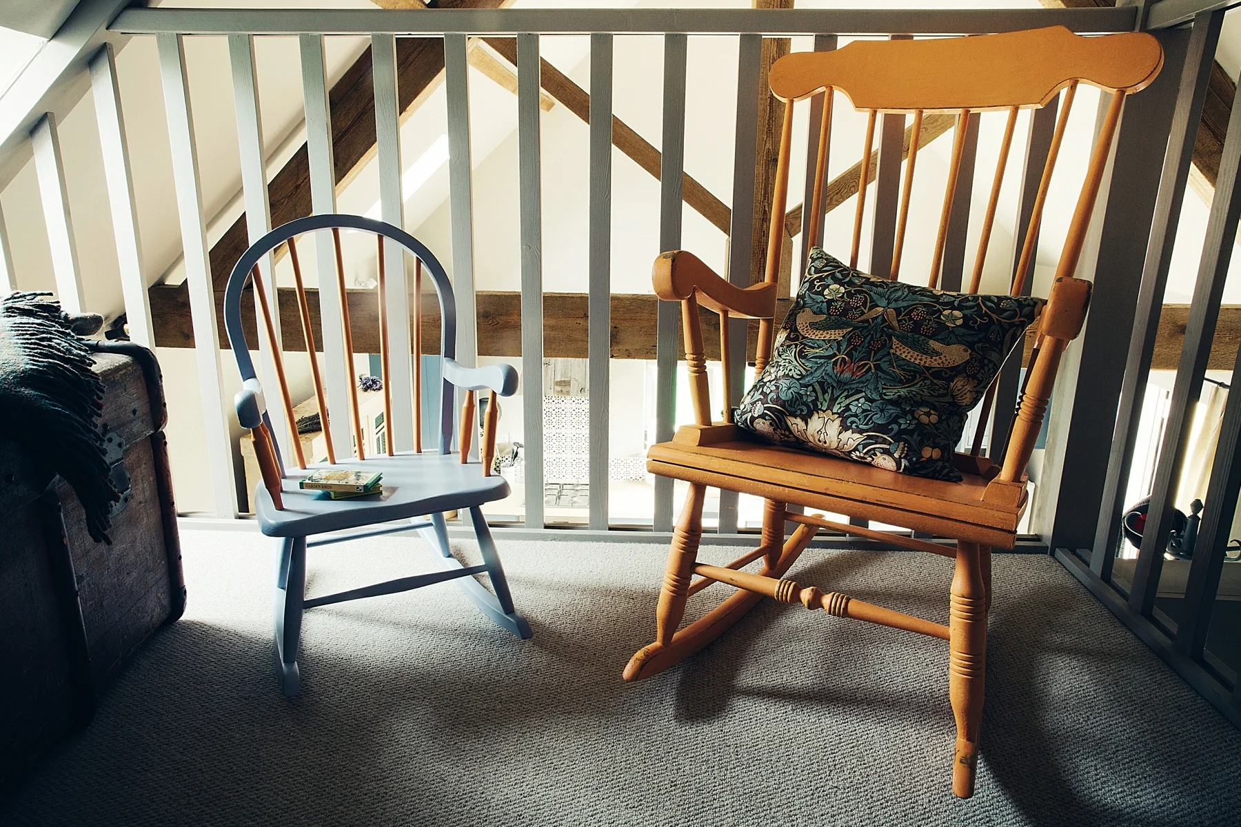A blue wooden rocking chair and a wooden armchair with a decorative pillow are placed on a carpeted floor in front of a staircase with vertical white railing and wooden support beams.