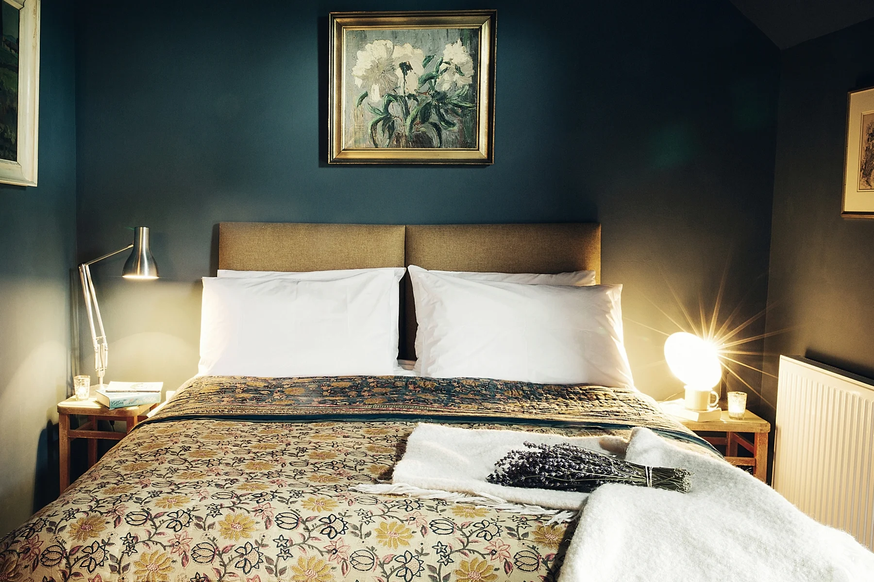 A neatly made bed with white pillows and a patterned bedspread in a cozy bedroom, with bedside tables, a lamp, and framed artwork on dark green walls.