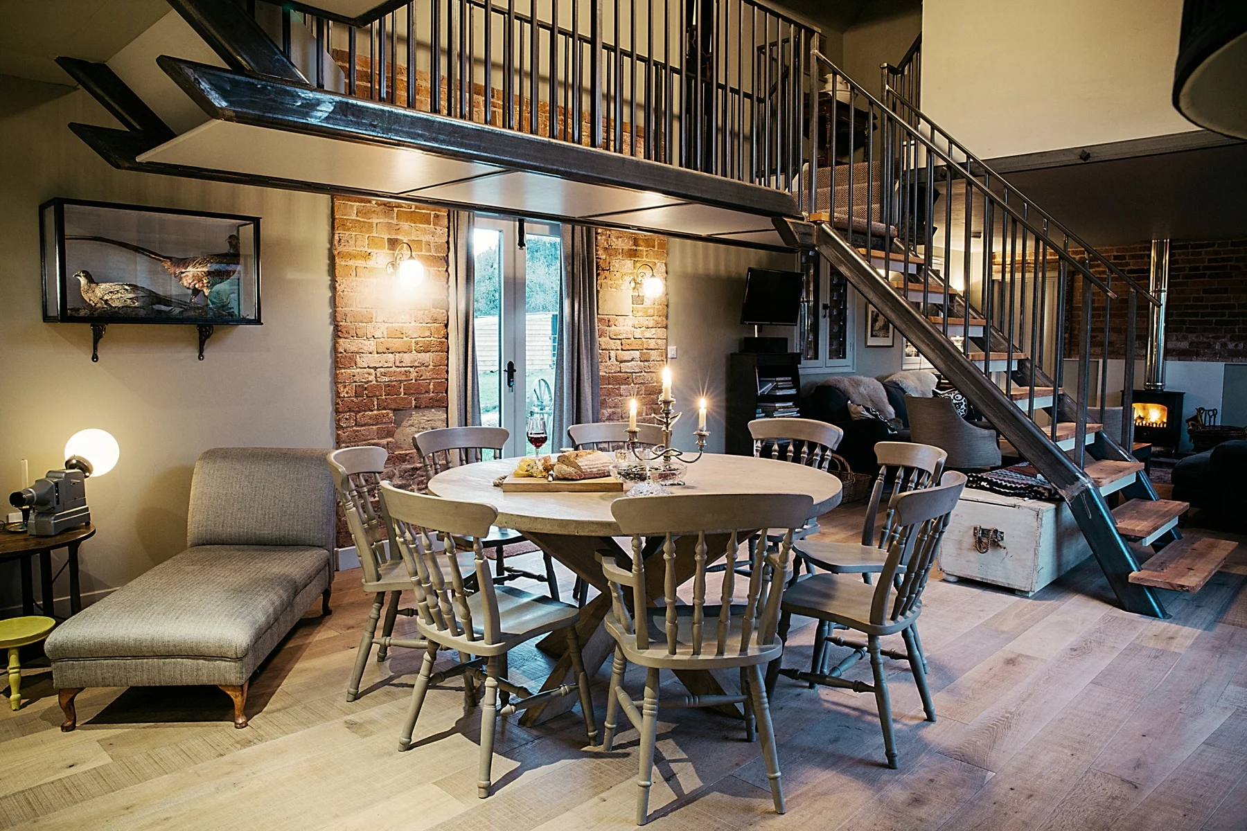 Living and dining area with brick wall, round dining table surrounded by wooden chairs, a daybed, and a staircase leading to a second level. There are candles on the table, a television, and decorative items including a painting of birds on the wall.