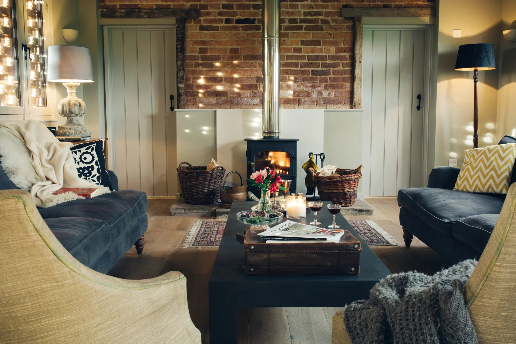 Living room with a brick fireplace, two sofas, a central coffee table with candles, wine glasses, and a flower arrangement, and various lamps and decor items.