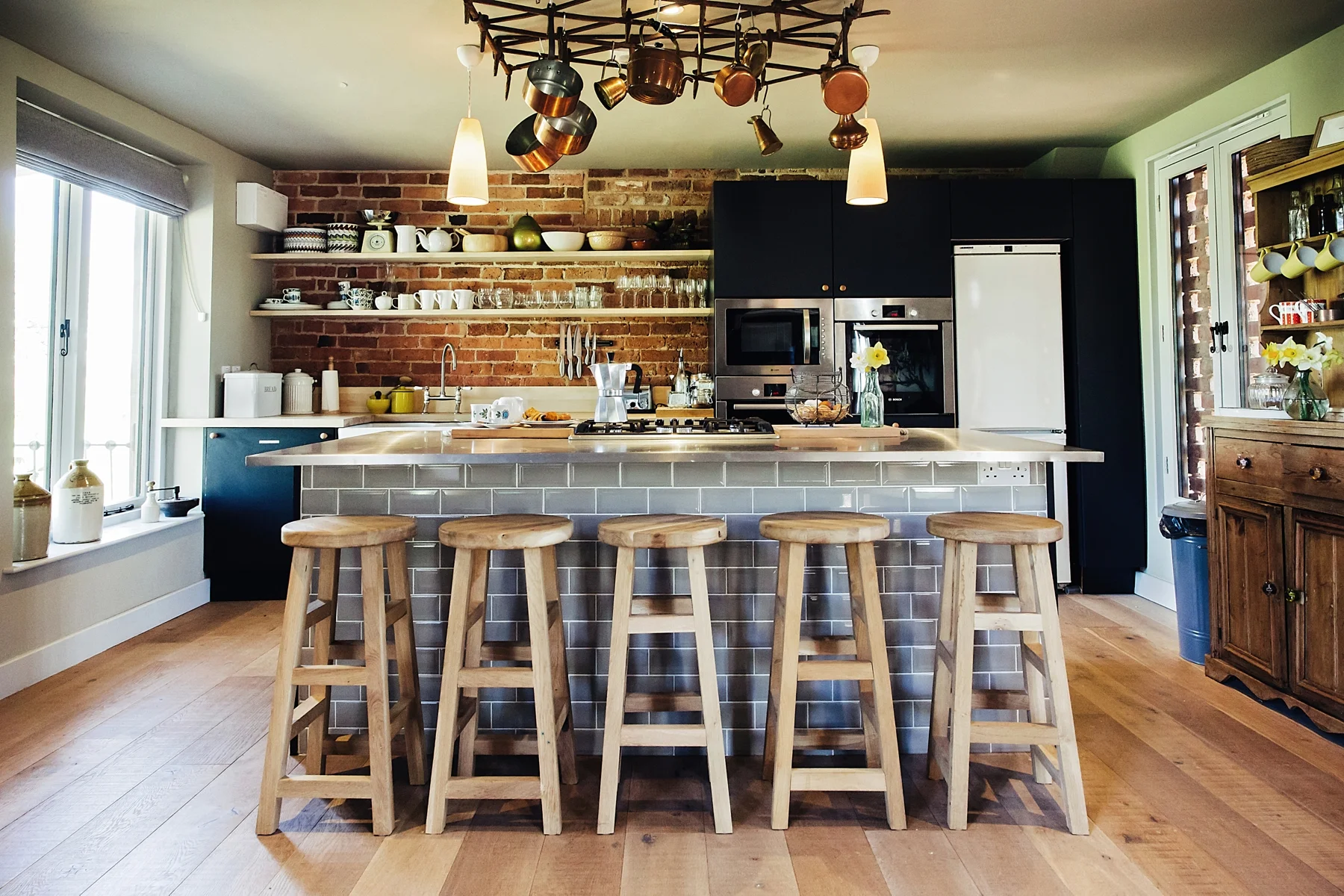 Modern kitchen of Redford Farm Barns with a brick wall, open shelves with dishes, a kitchen island with five wooden stools, and stainless steel appliances.