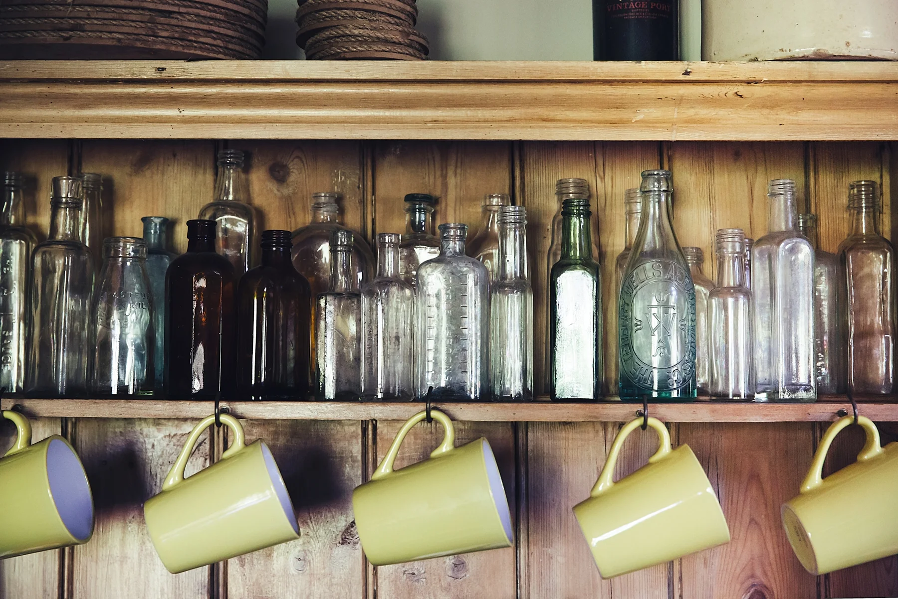 A wooden shelf holding empty glass bottles of various shapes and sizes, with four yellow coffee mugs hanging underneath.