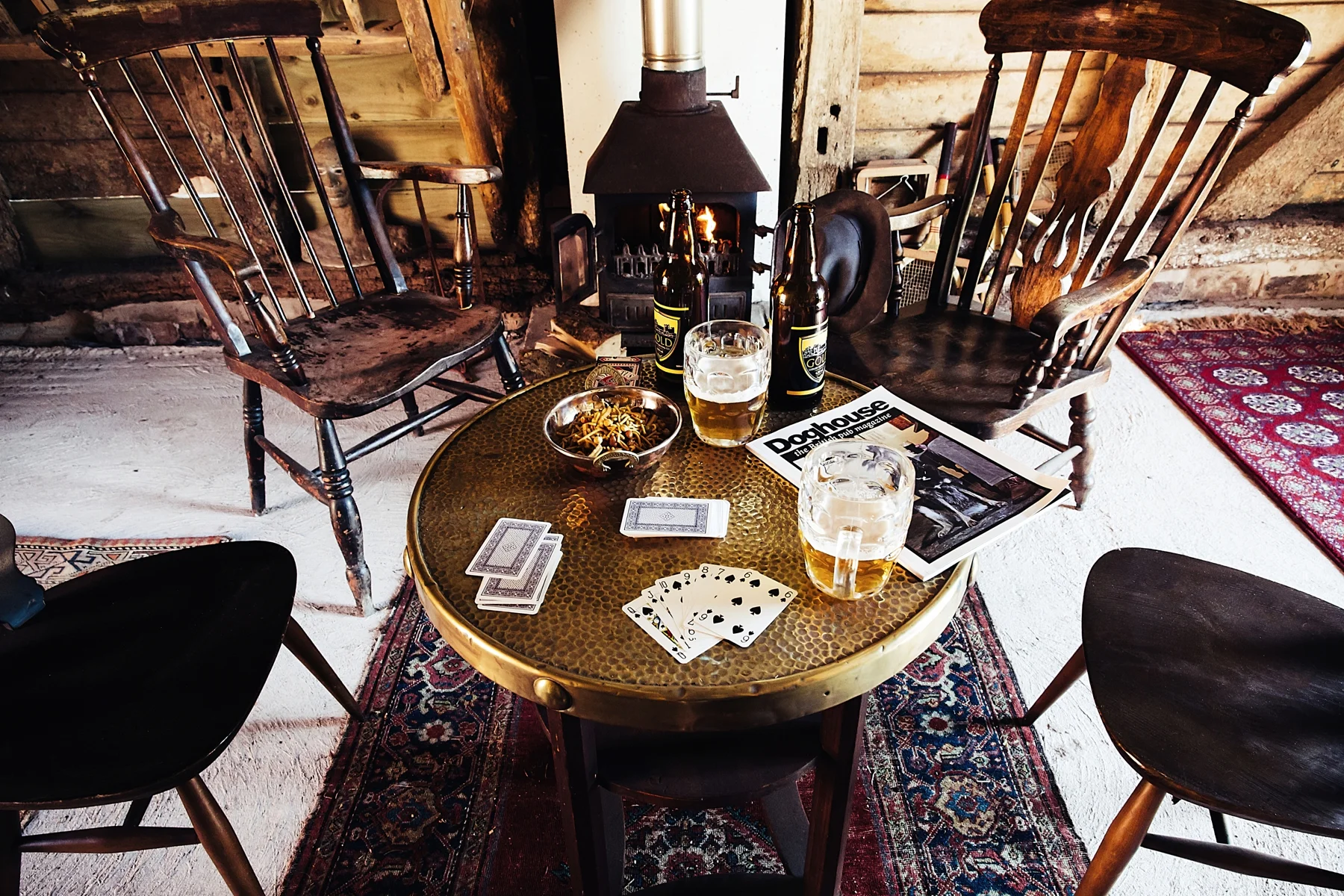 A round table with a hammered brass surface holding a deck of cards, two glasses of beer, a bowl of snacks, and a magazine titled "Dachouse" sits in front of a fireplace. Four wooden chairs surround the table in a rustic setting with wood-paneled wal