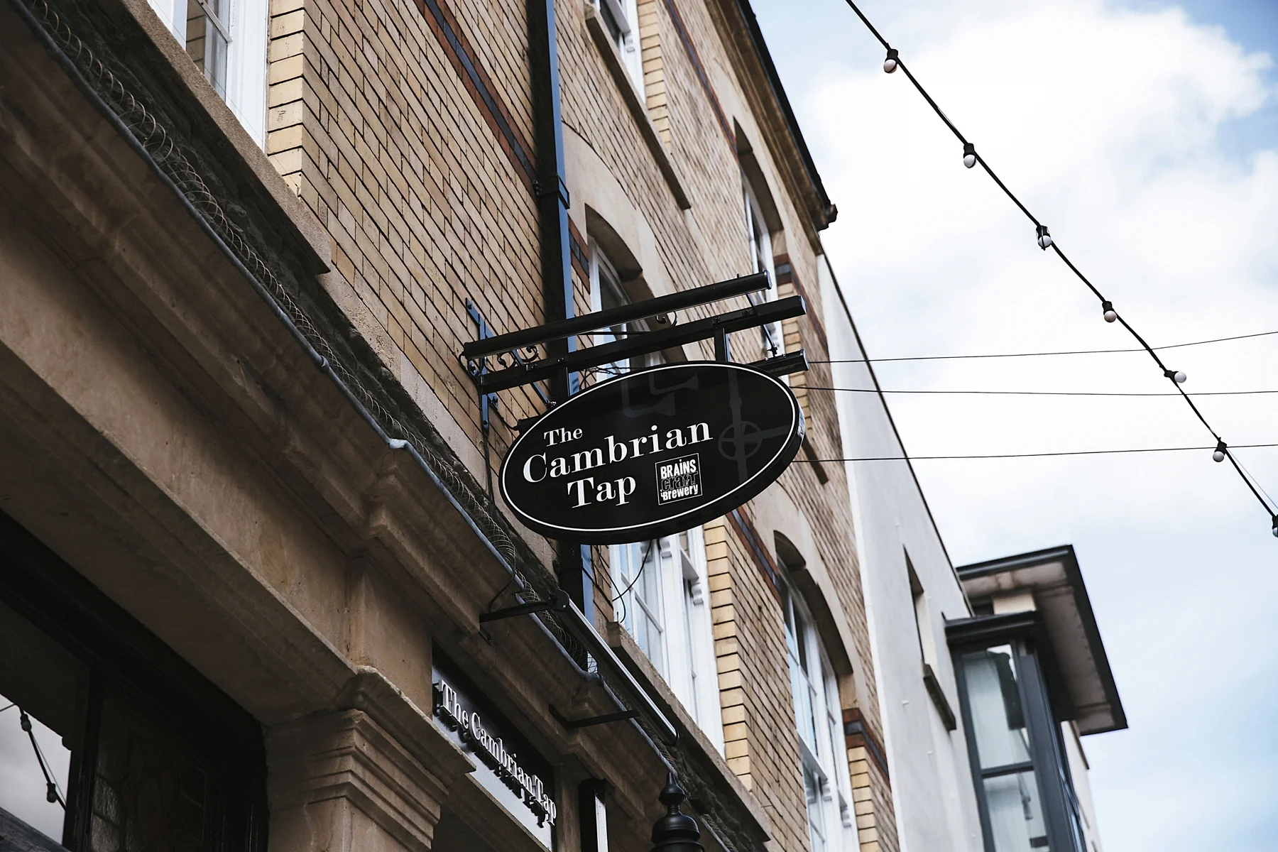 Sign for The Cambrian Tap brewery hanging outside a brick building with string lights above.