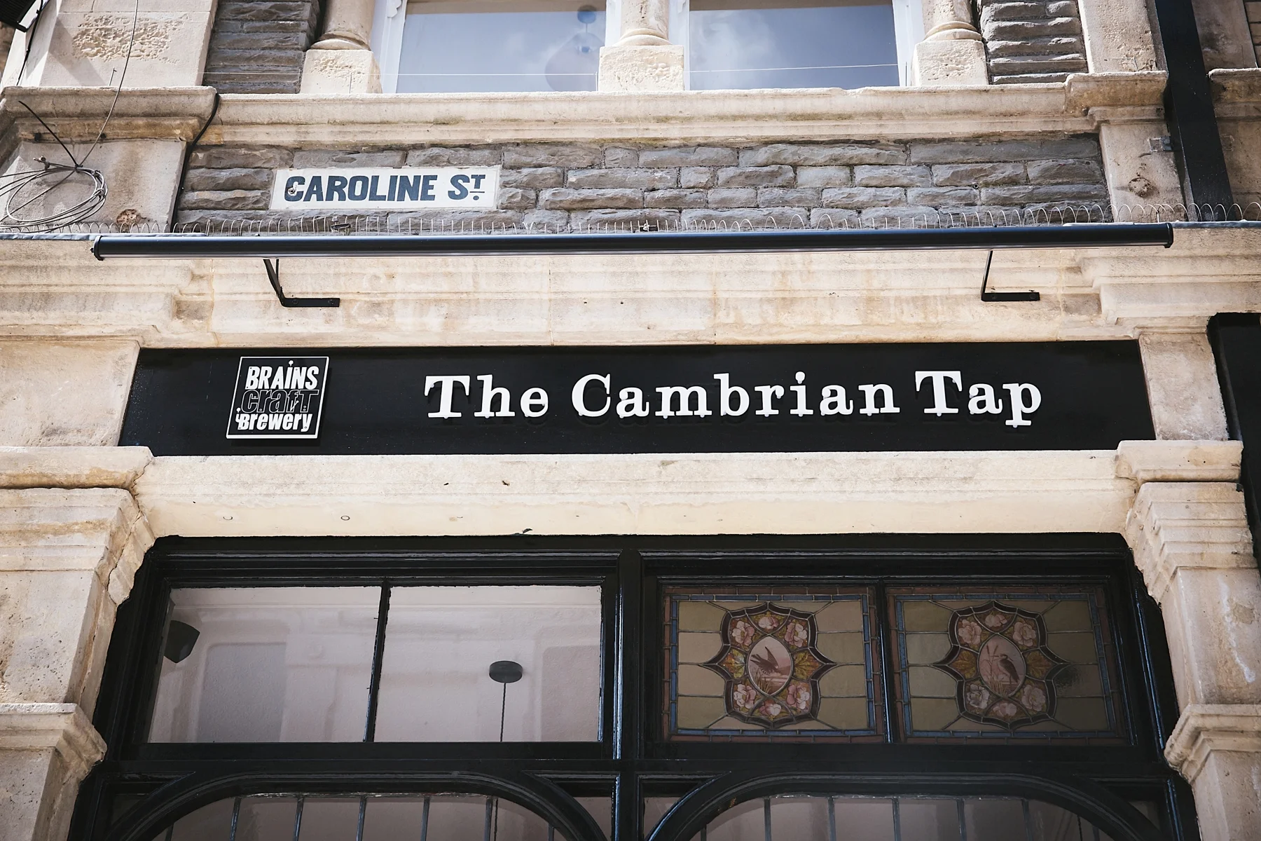 Storefront signage for The Cambrian Tap, part of Brains Craft Brewery, on a stone building with a second-floor window and street sign for Caroline Street.