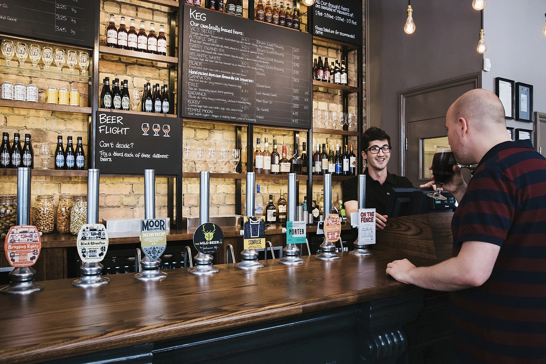 A man with a shaved head wearing a striped shirt is at a bar holding a glass of dark beer. Behind the bar, a bartender is smiling. The bar has a wooden counter with beer taps displaying various labels and a brick wall with bottles, glasses, and a cha