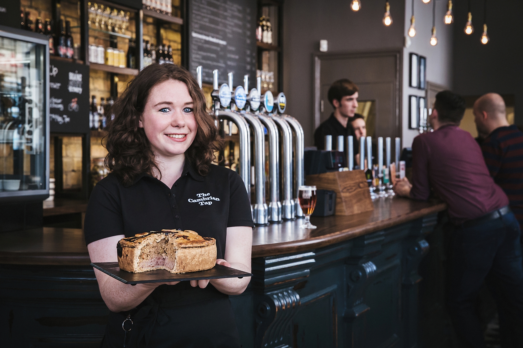 A smiling woman wearing a black shirt with the logo 'The Cambrian Tap' holds a black plate with a sliced fruit tart in a bar setting. Behind her are bar taps, drinks, and people sitting and standing at the bar.
