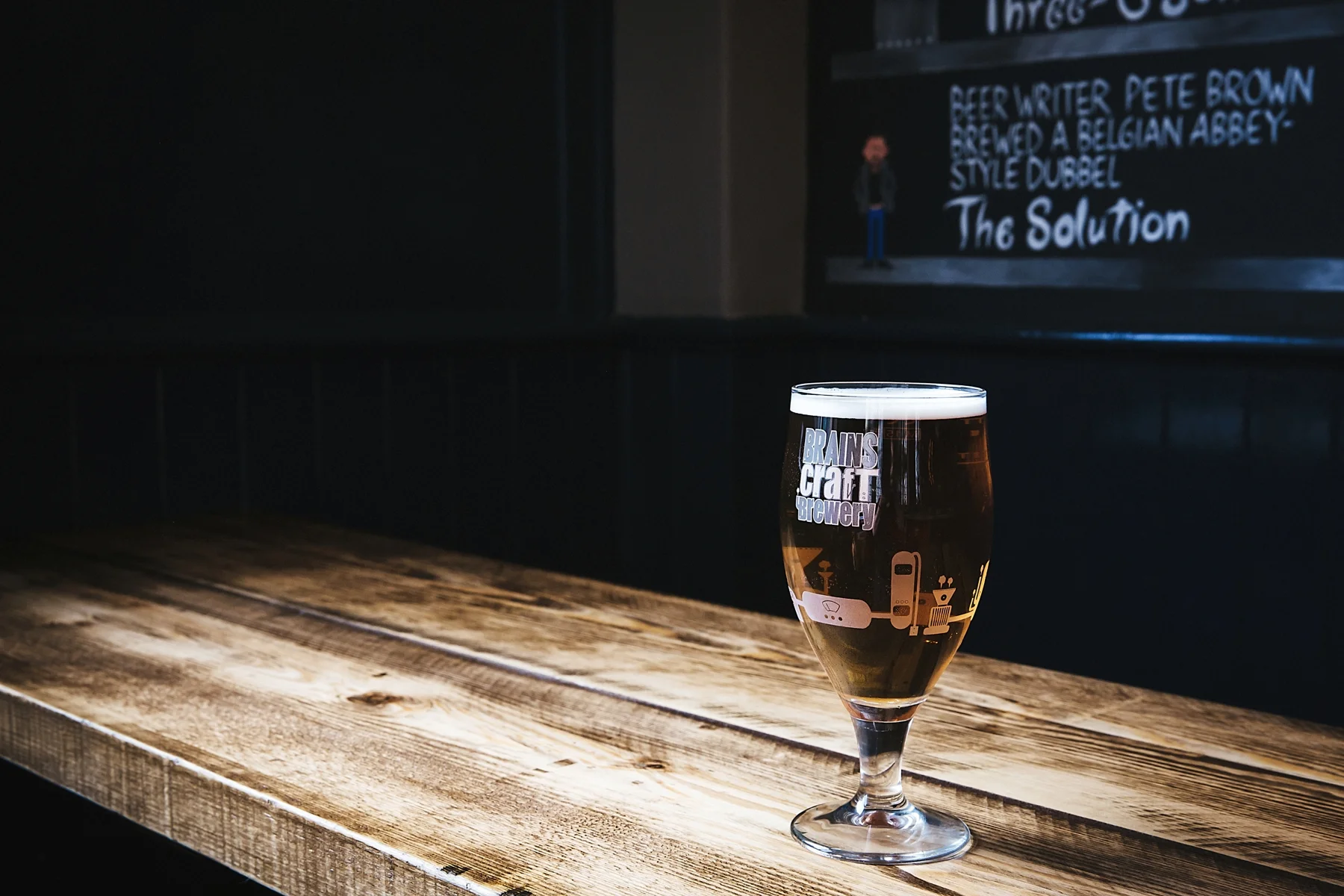 A glass of beer on a wooden table with a brewery logo, in front of a chalkboard menu at a bar.