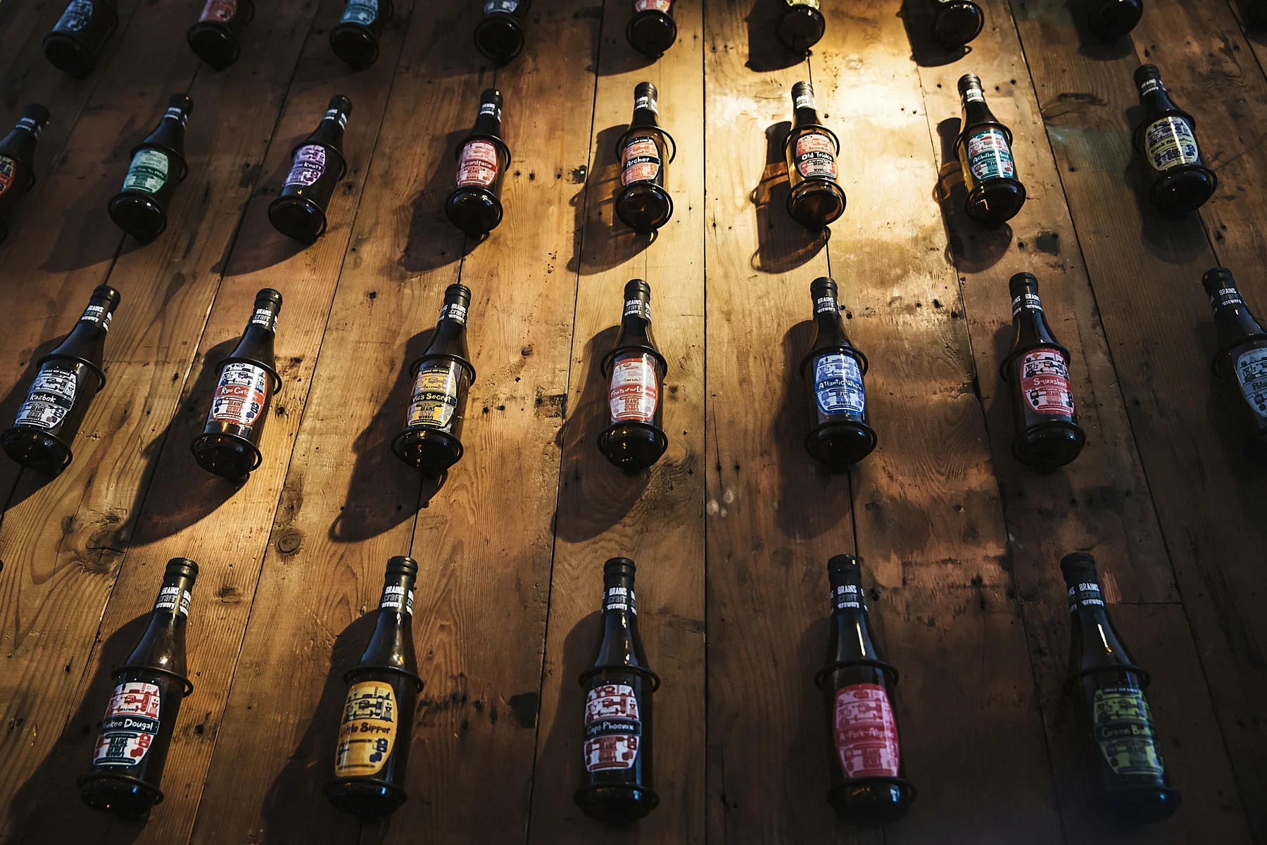 Bottles of craft beer with colorful labels arranged on a wooden wall, casting shadows.