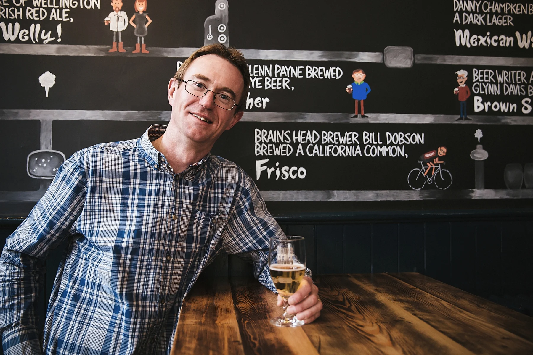 A man with glasses in a plaid shirt is sitting at a wooden table holding a glass of beer, smiling at the camera. Behind him is a blackboard with colorful drawings and text about various beers and their descriptions.