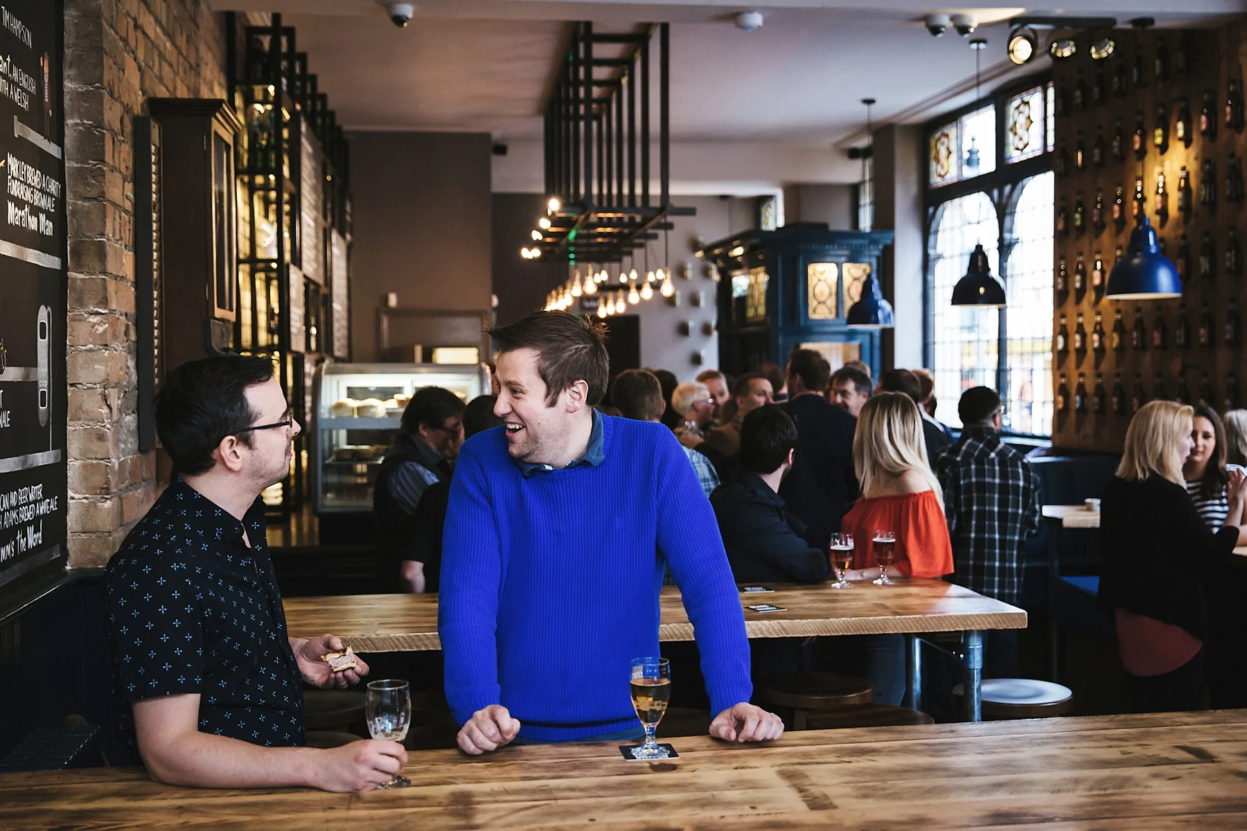 Two men are talking and smiling at a bar in a lively restaurant, with other patrons in the background.
