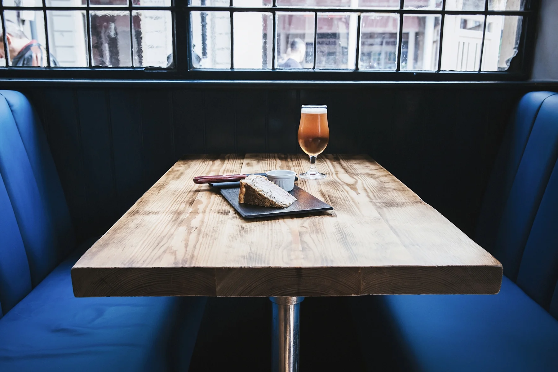A wooden table with a slice of cake, a small bowl of sauce, and a glass of beer inside a restaurant with large windows and blue cushioned seating.