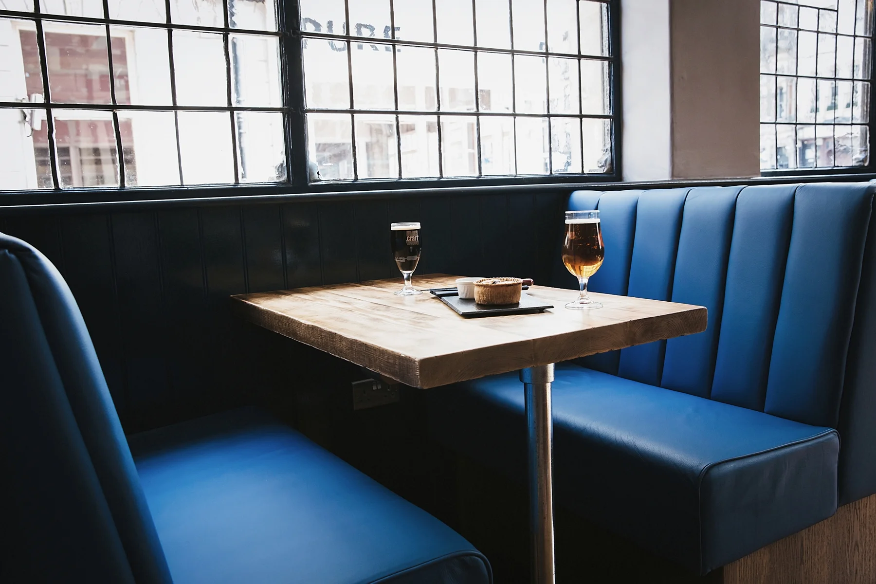 A wooden table with two glasses of beer, a small bowl, and a napkin holder inside a restaurant with blue upholstered booth seating and large window with black framing.