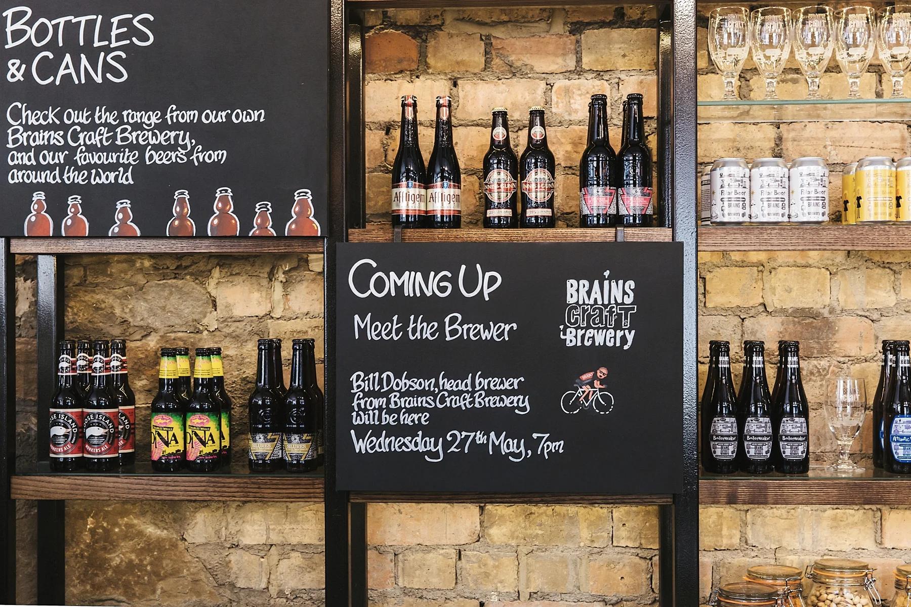 Shelves displaying bottles of craft beer, glasses, and cans, with a chalkboard sign showing upcoming brewery event details at Brains Craft Brewery.