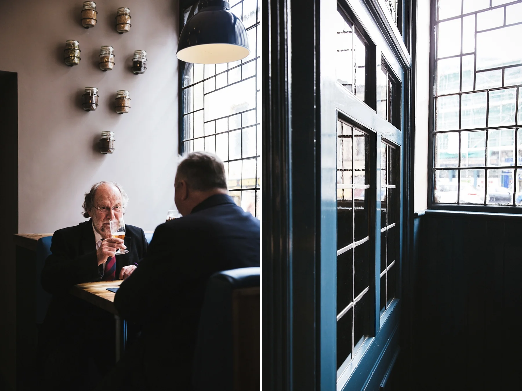 Two men in suits sitting at a table in a restaurant, drinking beer, with a large window and brick wall behind them, and shelves with jars on the wall.