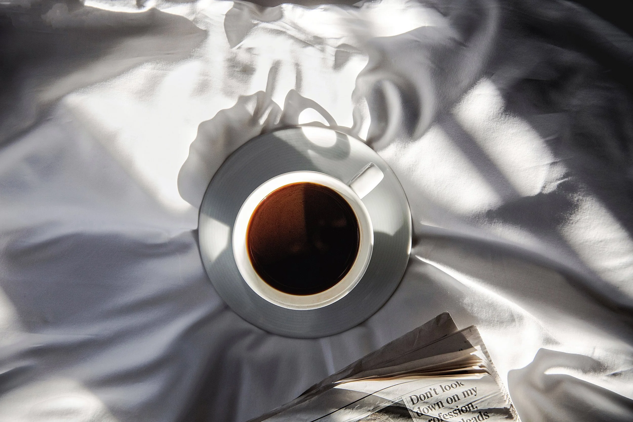 Top-down view of a white coffee cup filled with black coffee, placed on a white saucer with a silver rim, on a white bed with a person's hand resting nearby. A newspaper page with partial text is also visible at the bottom of the image.