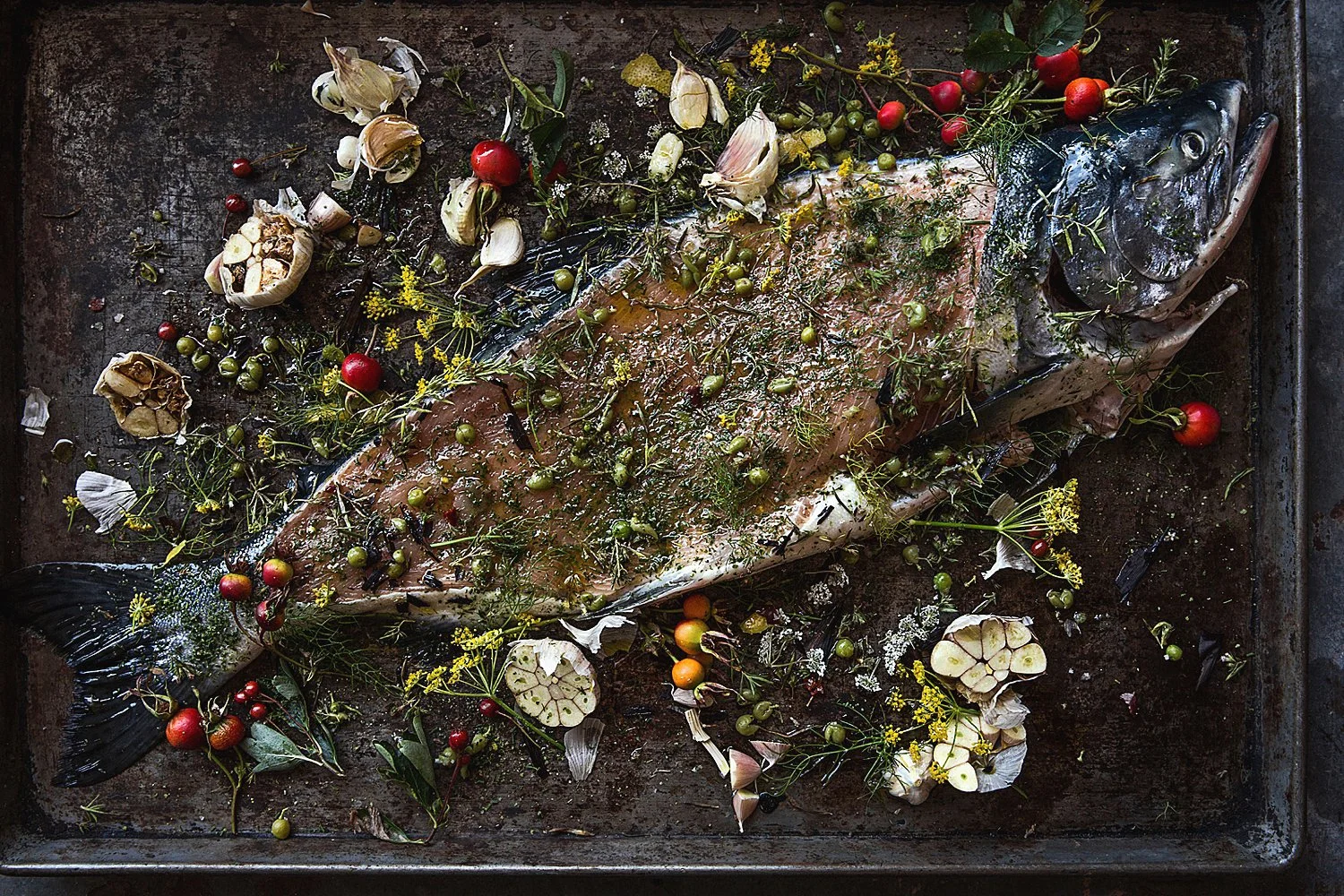 Raw fish on a tray garnished with herbs, cherry tomatoes, garlic bulbs, and vegetables.